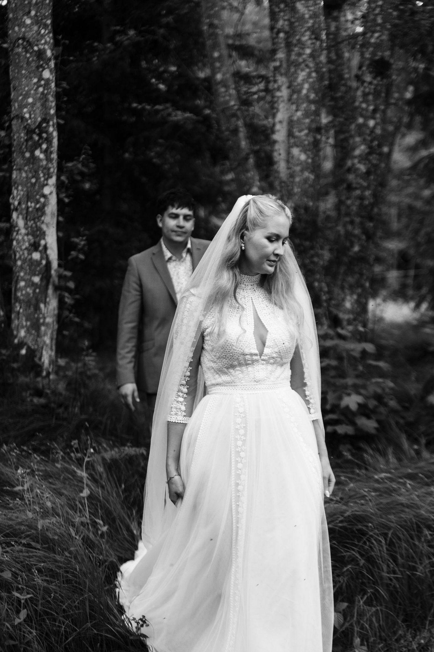 Black and white wedding photo of bride in veil and gown with groom in suit among forest trees.