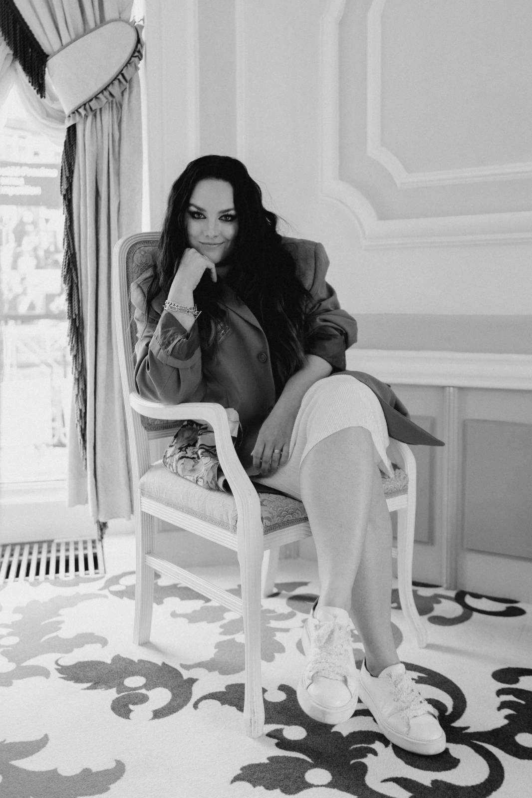 A person in a white dress sits on a chair in a bright room with ornate floor patterns and window light.