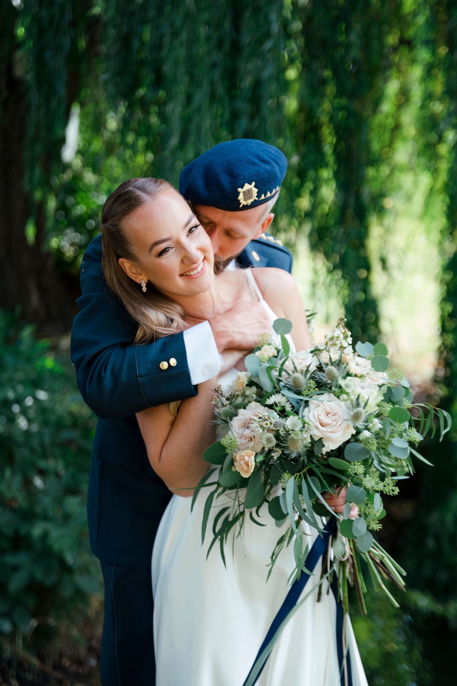 Wedding couple embracing under willow trees with elegant white and green floral bouquet.