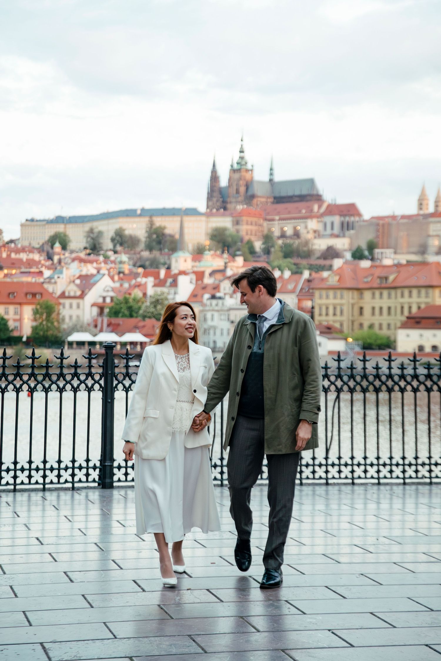 Couple walking along Prague Castle viewpoint with historic skyline in background.
