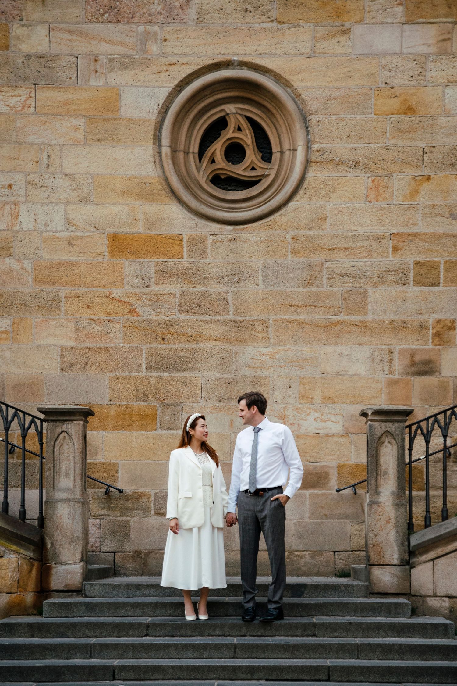 Couple stands on stone steps beneath circular window of historic building.