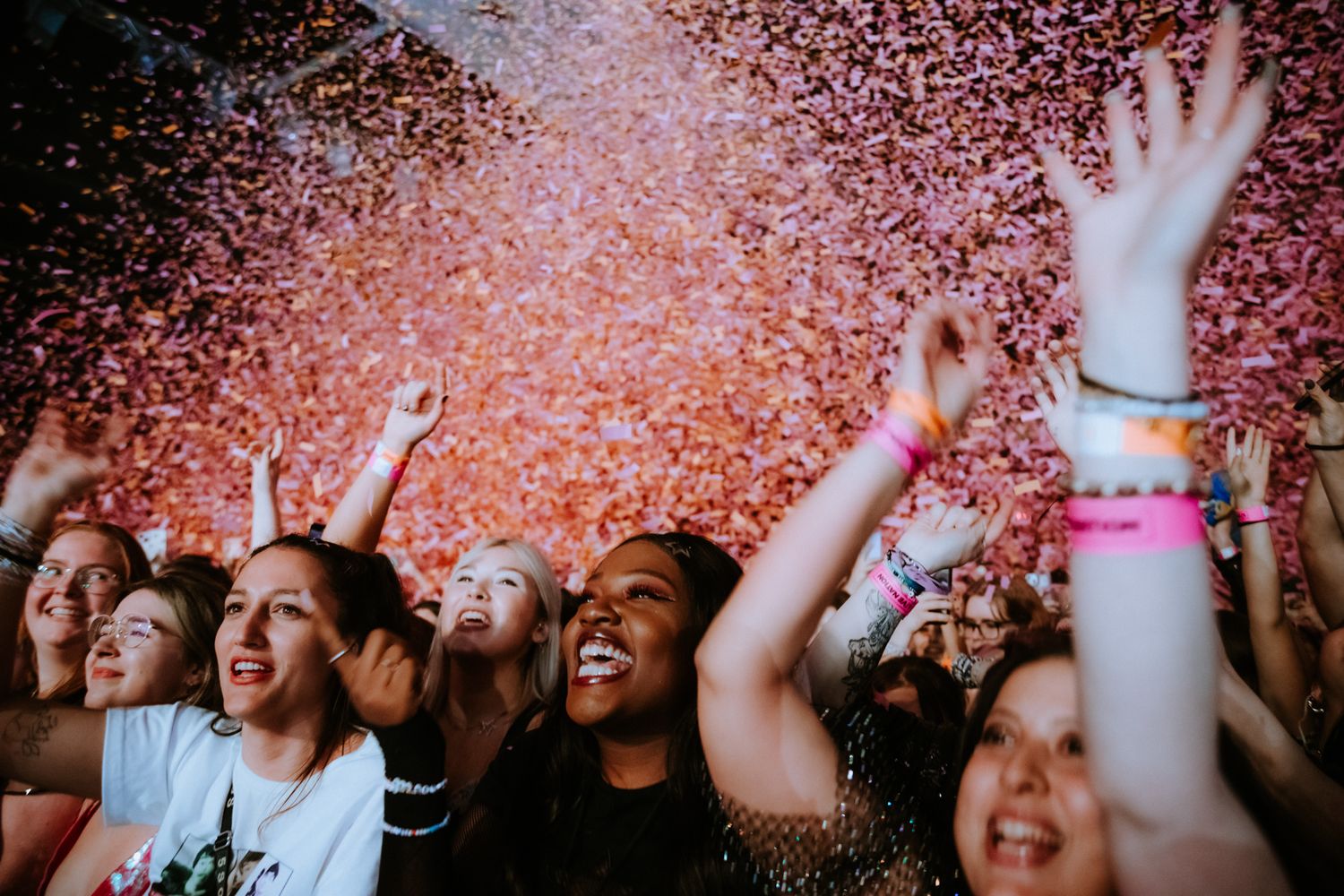 Excited concert crowd celebrates with hands raised while pink confetti rains down during a live music performance.