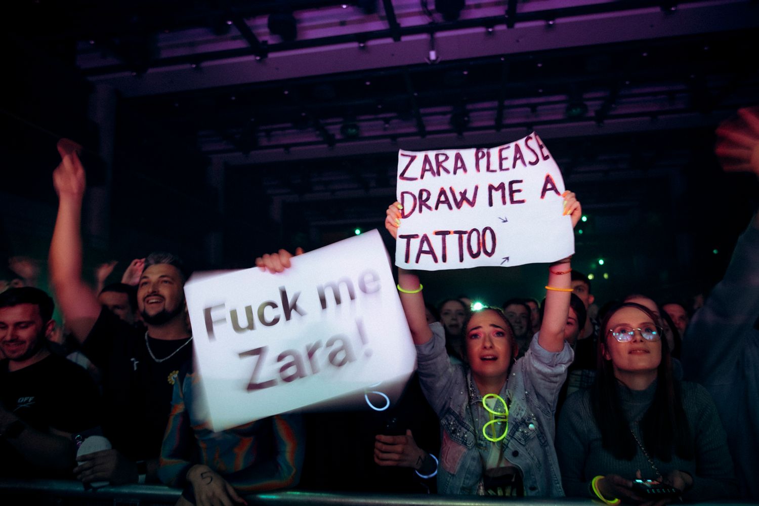 Concert crowd holds up handwritten signs during a performance in a dark venue with purple lighting.