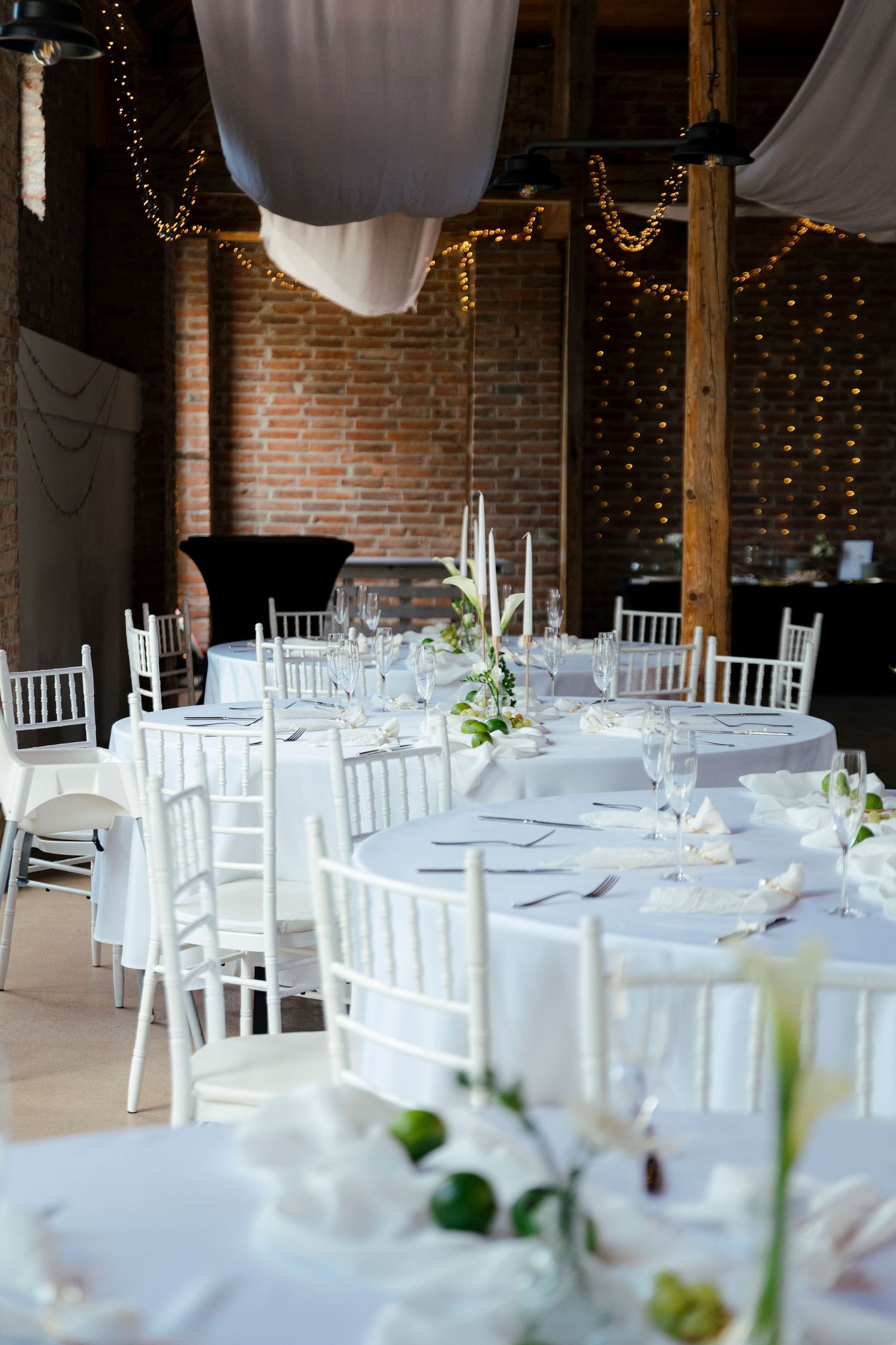 Elegant wedding reception in rustic brick venue with white round tables, chiavari chairs, candles, and fairy lights.
