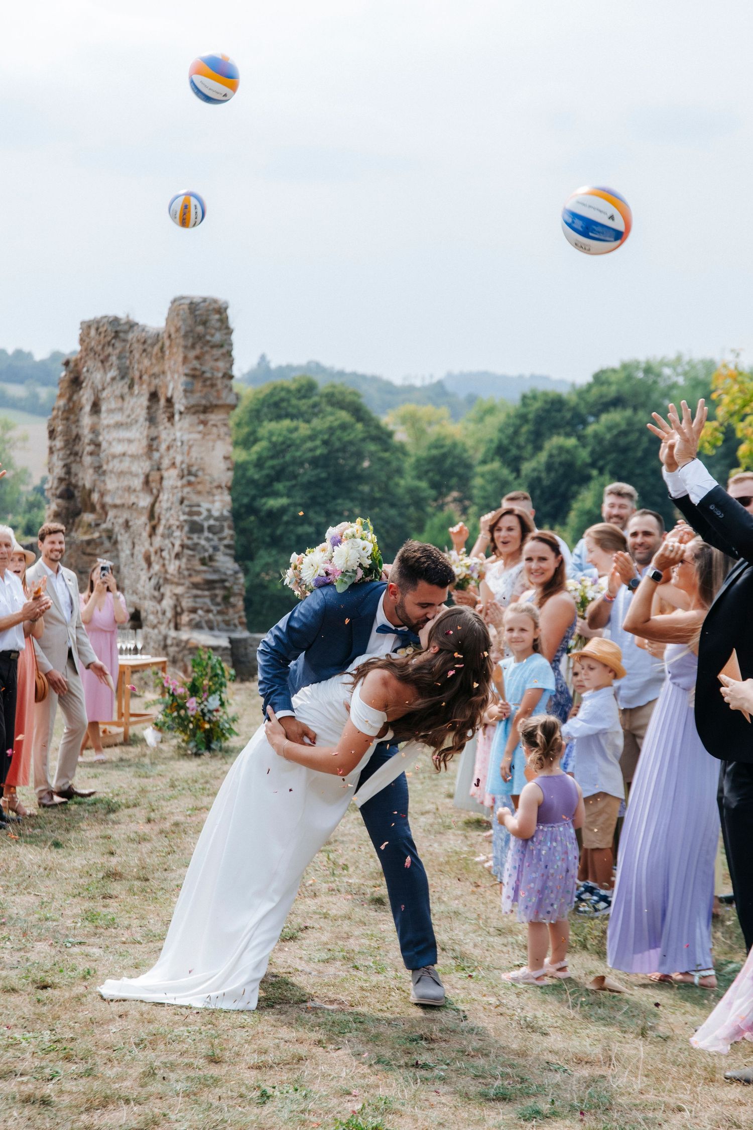Wedding guests toss beach balls in celebration near stone ruins during an outdoor summer ceremony.