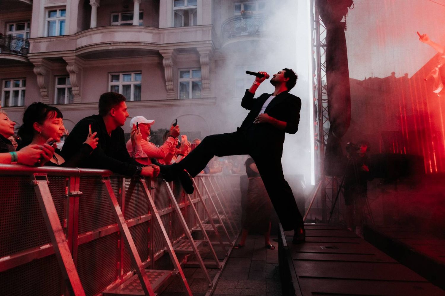 Performer on stage silhouetted against red smoke while singing at an energetic concert with crowd barriers.