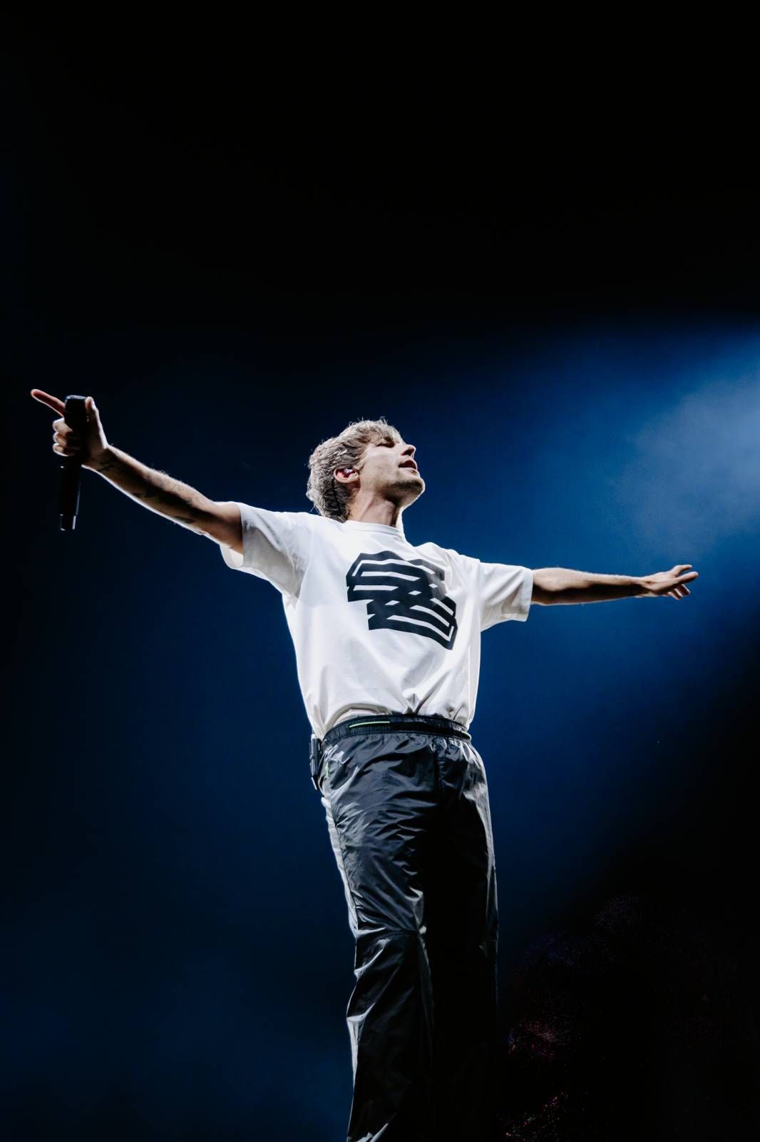A performer in a white t-shirt and dark pants strikes a dramatic pose with outstretched arms against a dark blue background.