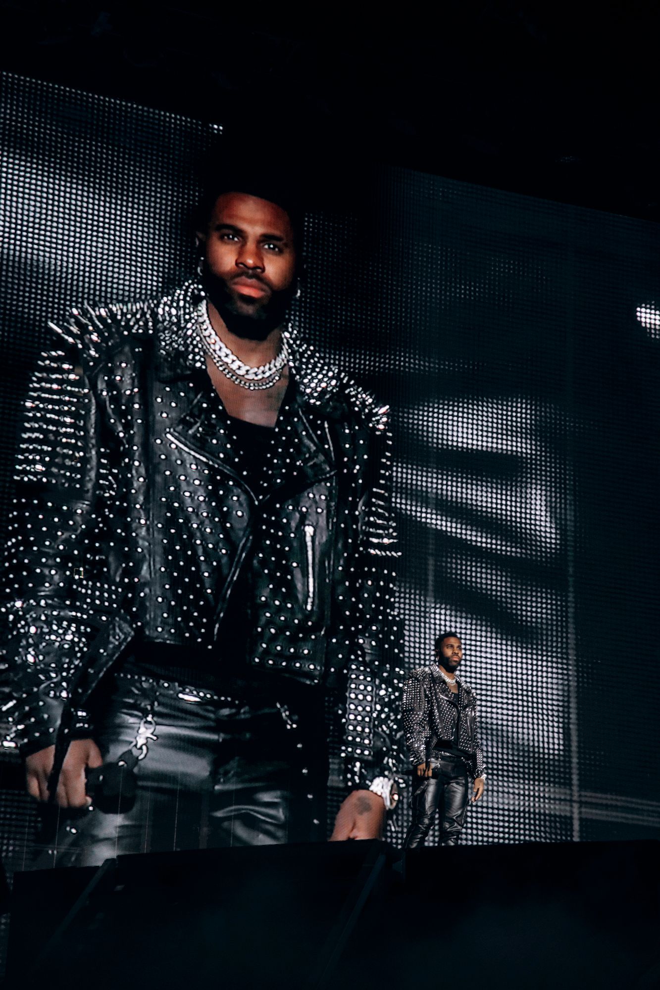 Performer wearing studded black leather jacket and chains performs on stage with dramatic lighting and video screens.