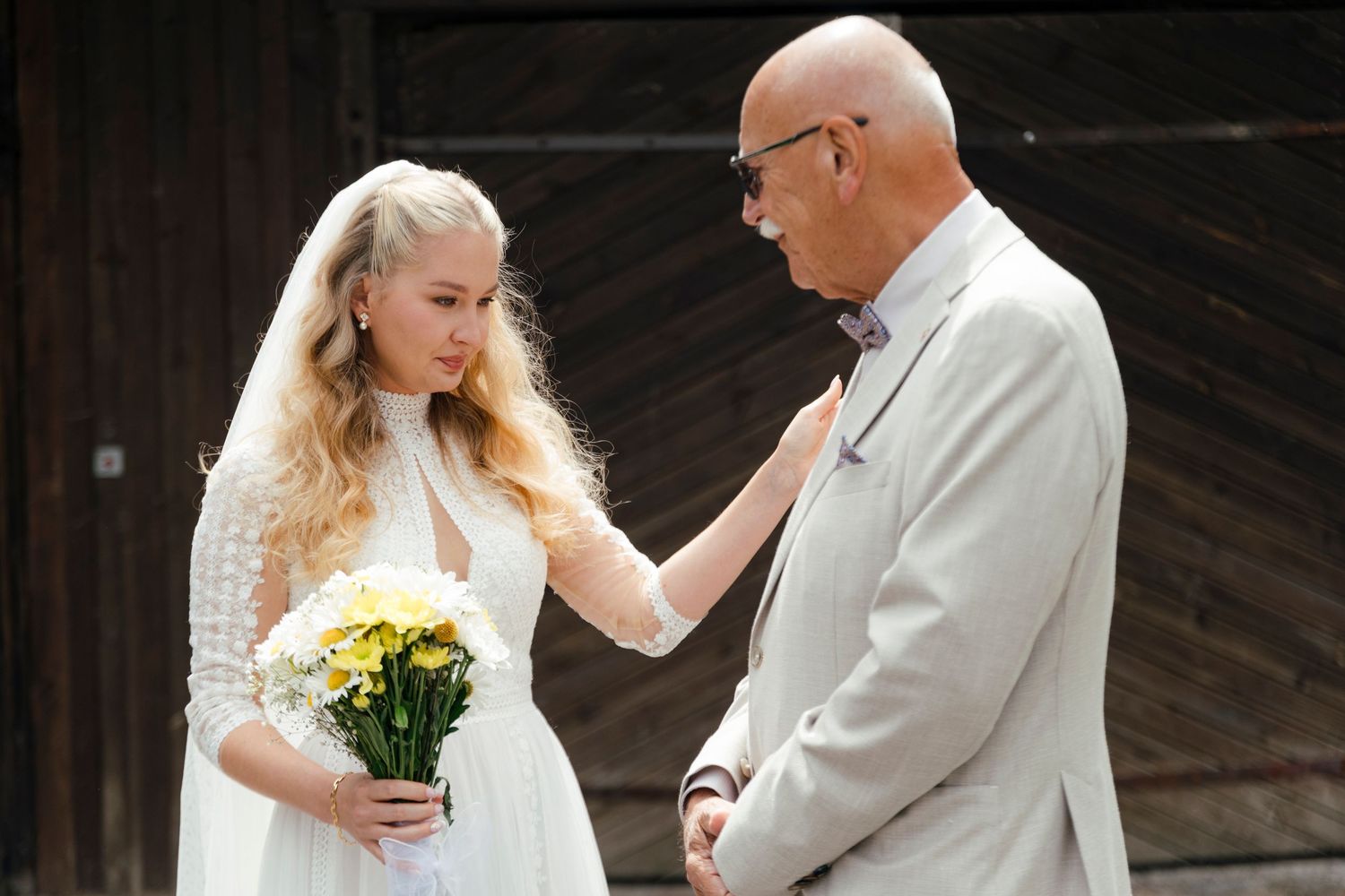 Bride in white lace dress holding yellow flowers adjusts groom's tie at outdoor wedding ceremony.