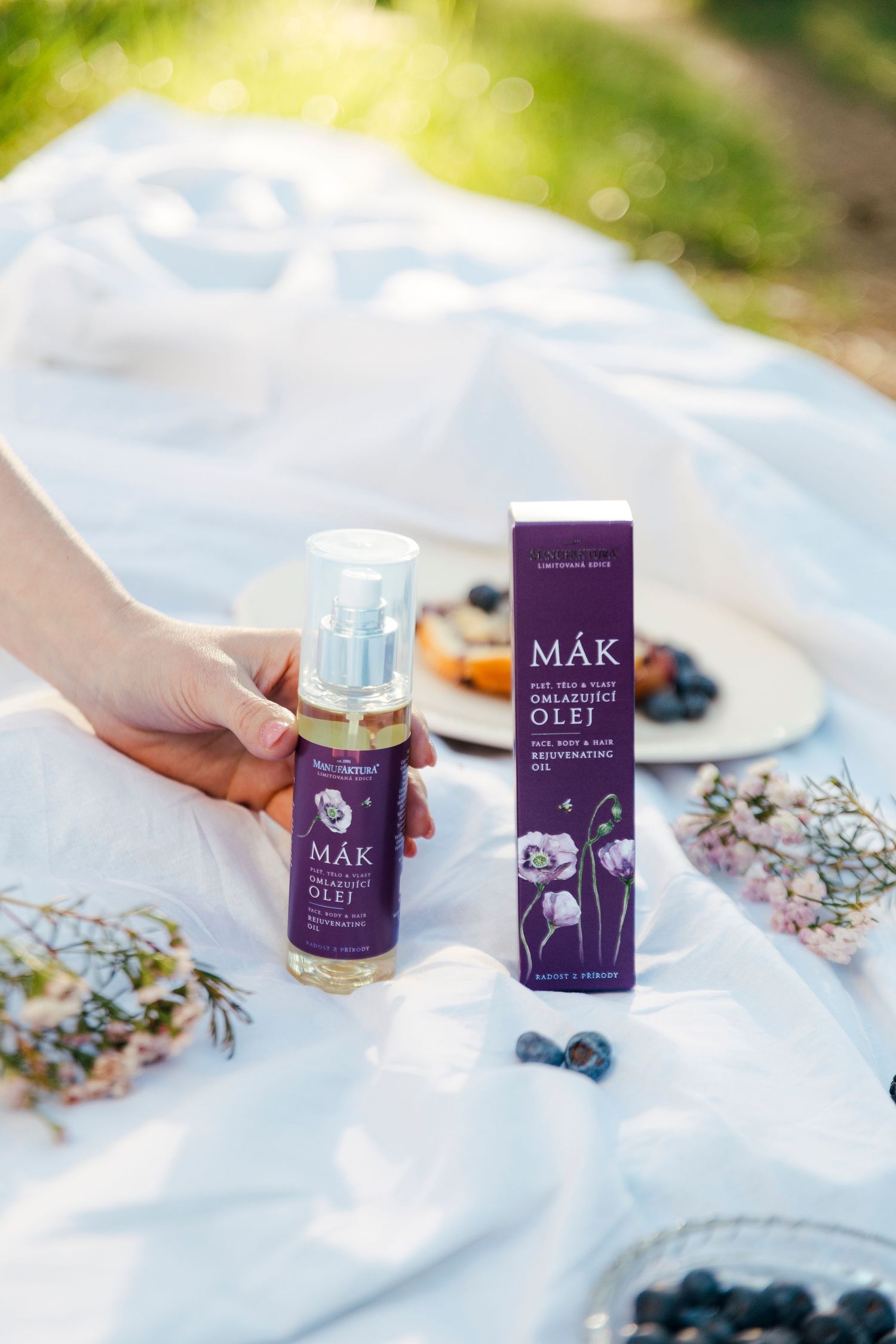Hand holding Mak face oil bottle beside purple box on white picnic blanket with blueberries and flowers.