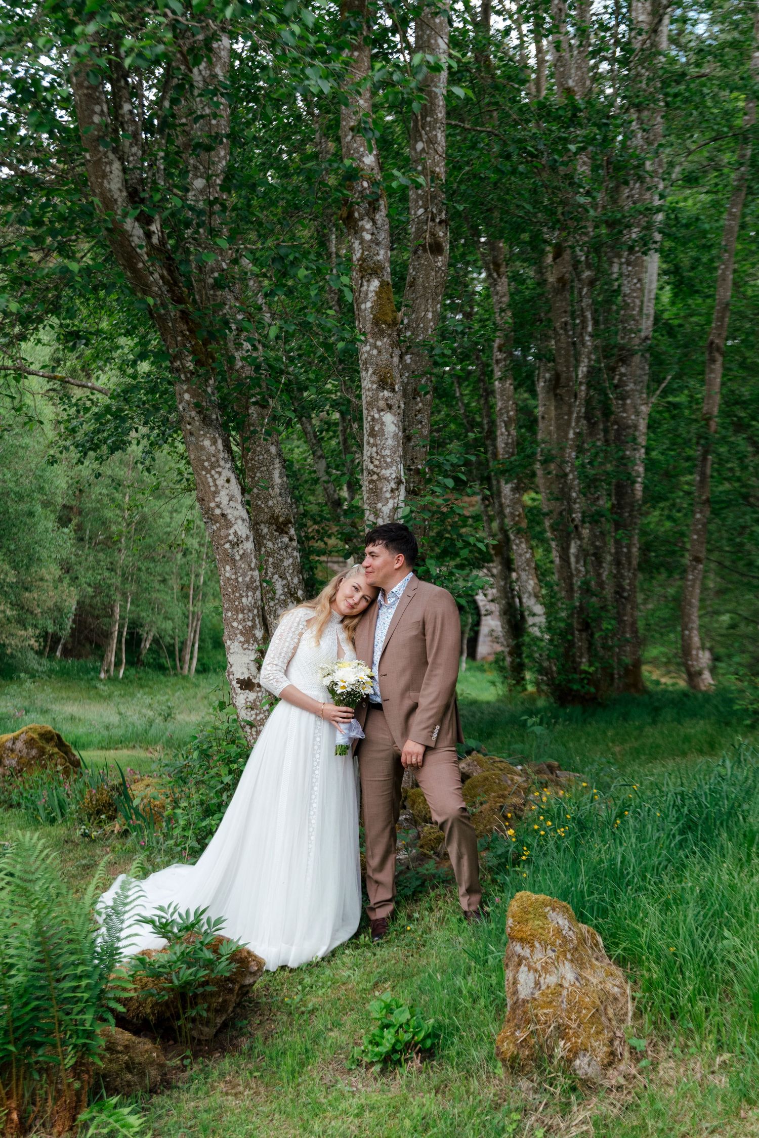 Bride and groom share a romantic moment in a lush green forest, with the bride holding a white bouquet.
