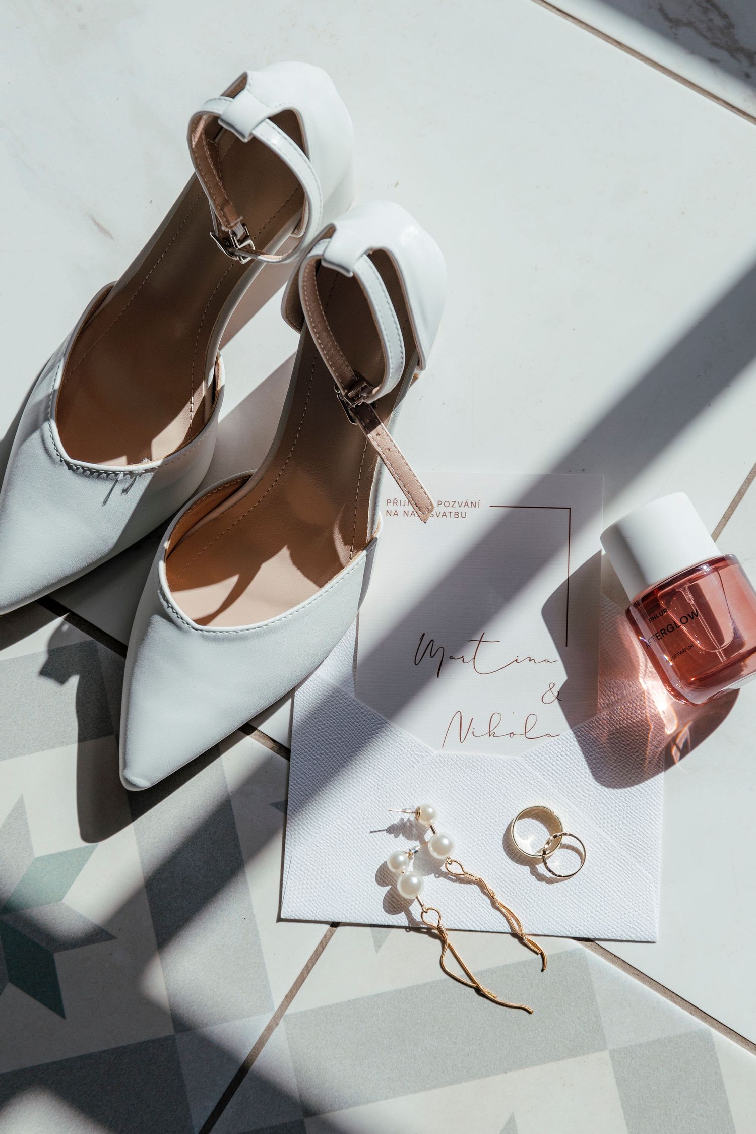 White bridal heels with pearl earrings, gold rings, pink perfume, and wedding invitation on marble tile floor.