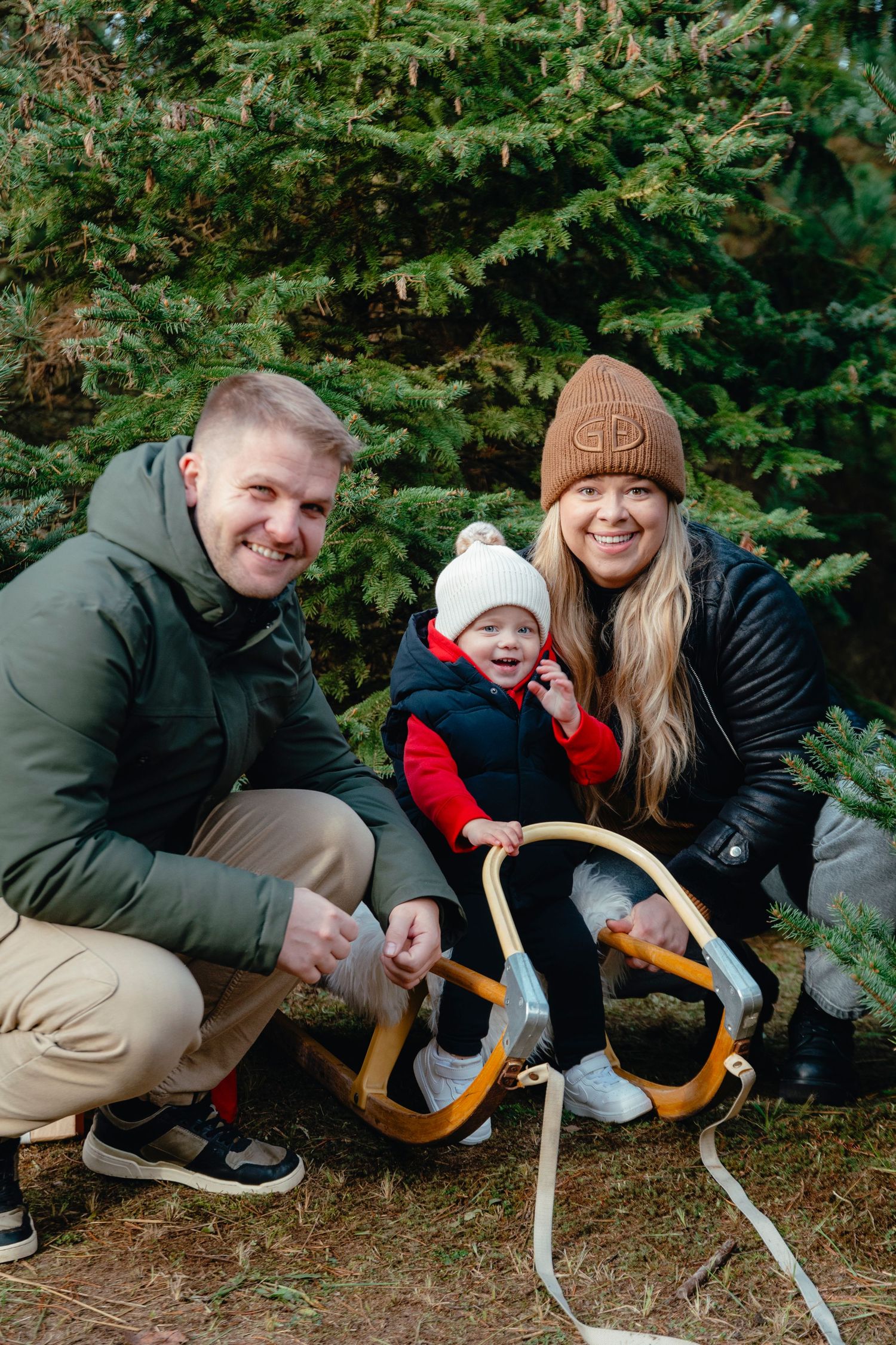 Family of three posing with a wooden sled near evergreen trees at a Christmas tree farm in winter.