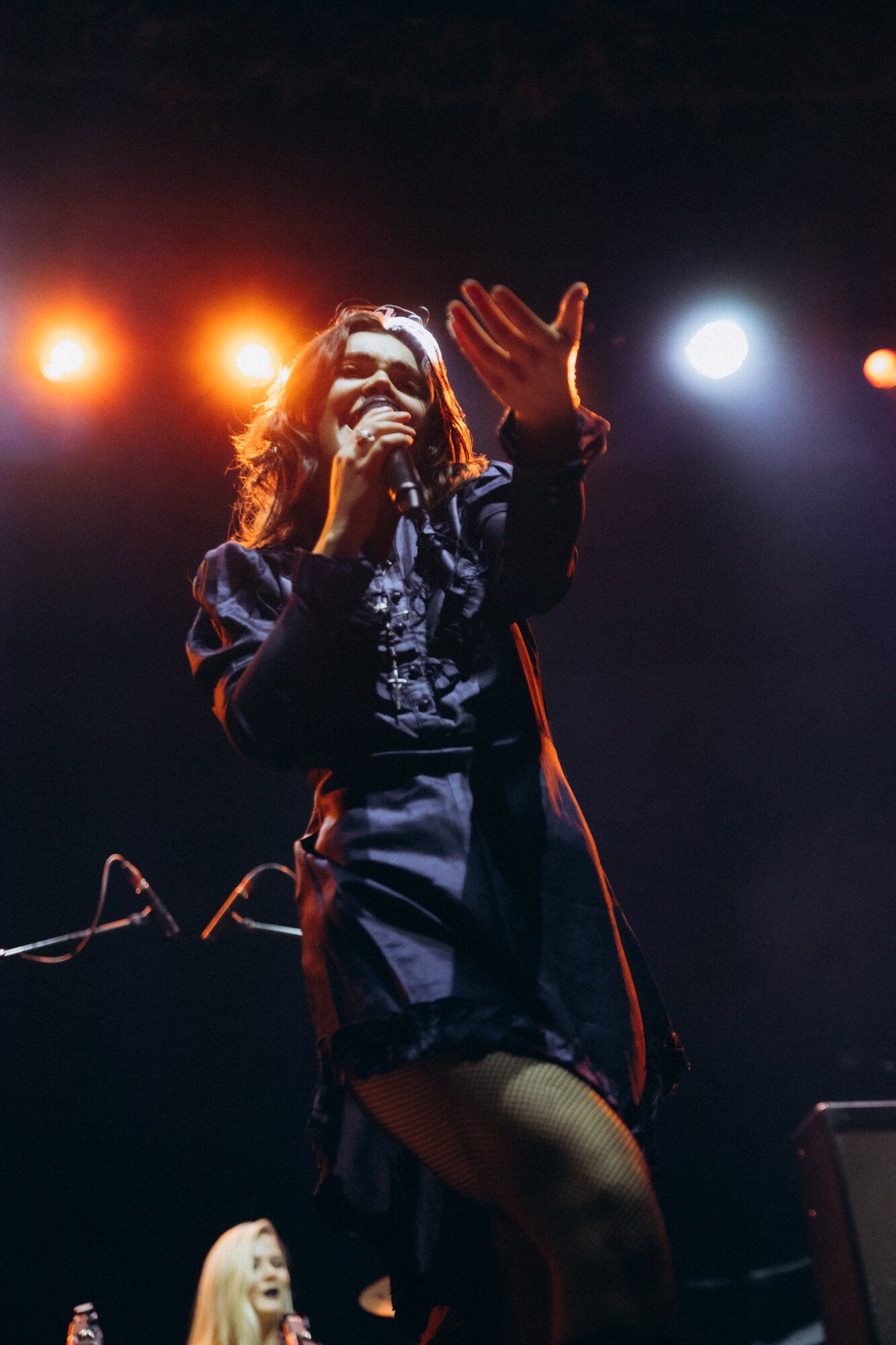 Performer in black outfit singing passionately on stage with dramatic orange and white stage lighting in background.