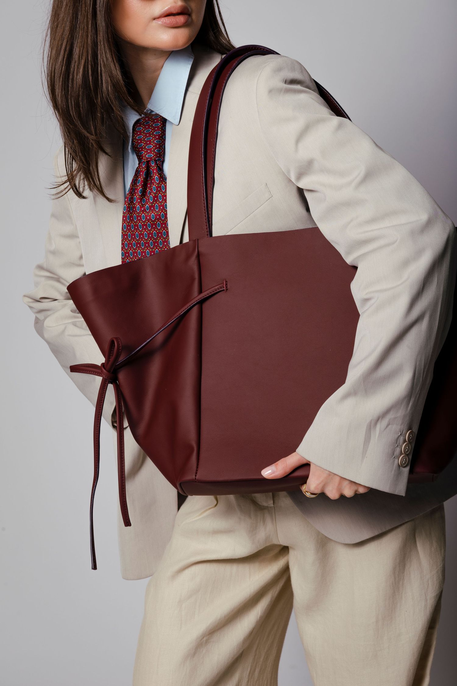 Burgundy leather tote bag with bow detail shown with beige outfit.