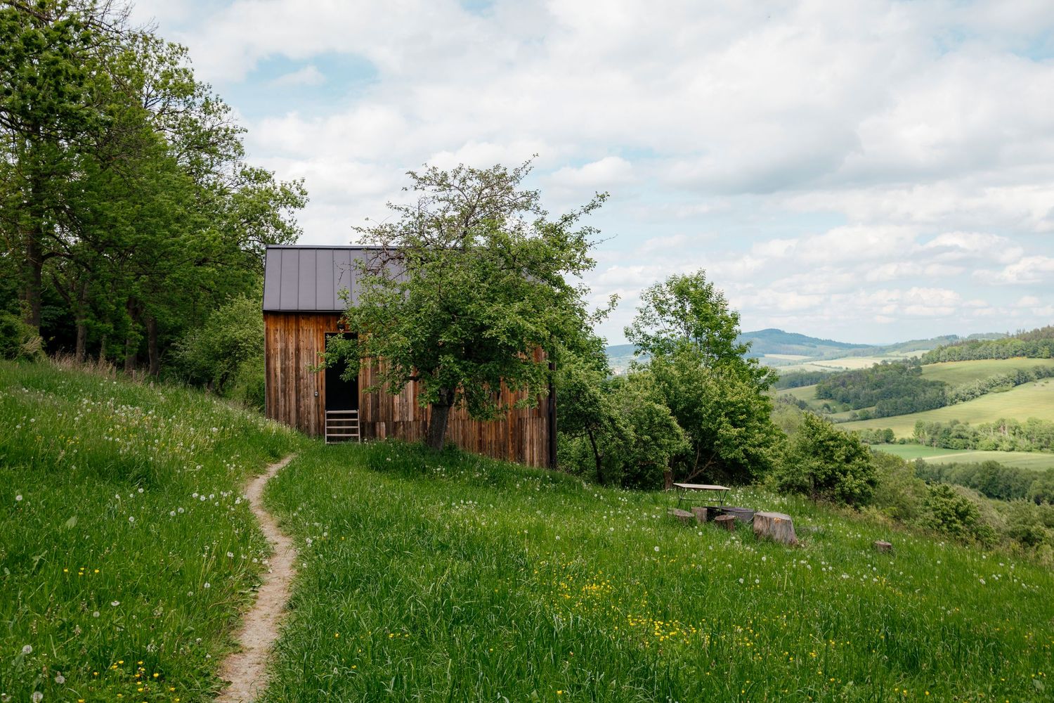 Rustic wooden cabin on a green hillside with mountain views and a winding path leading to the entrance.