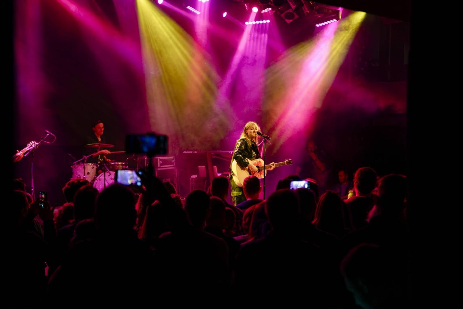 Concert performer on stage bathed in dramatic purple and yellow spotlights while audience members hold up phones.