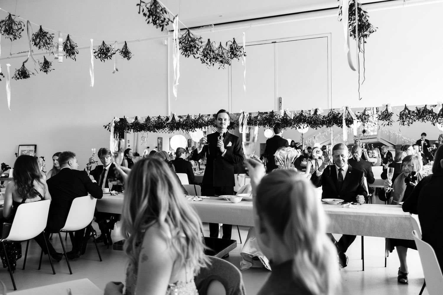 Wedding reception in a white-decorated banquet hall with hanging floral arrangements and seated guests.