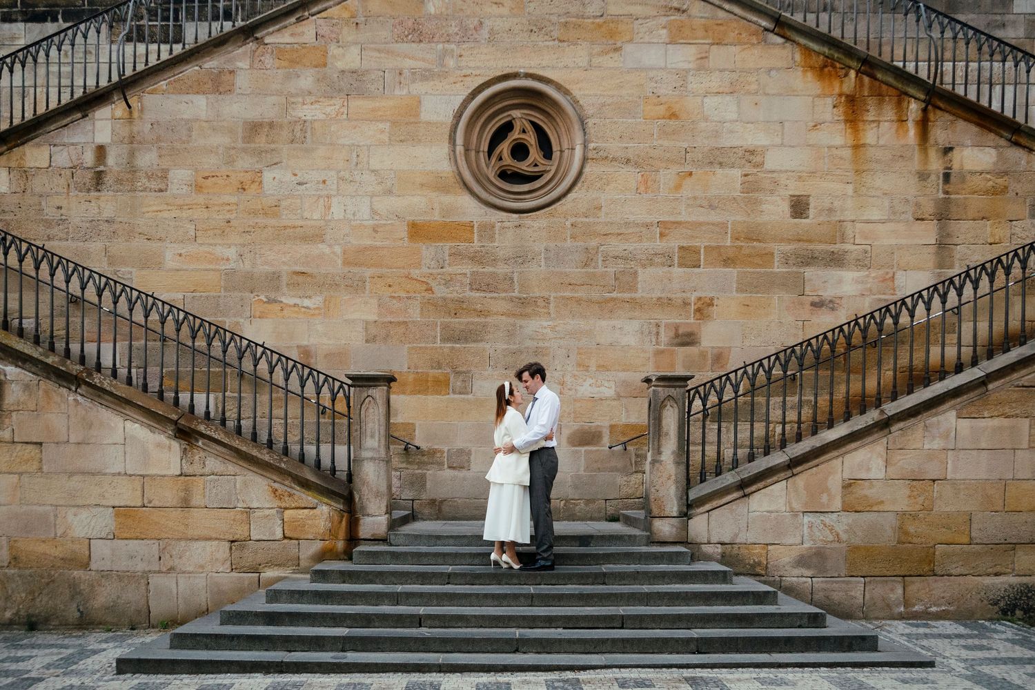 A couple poses on grand stone steps of a historic building with ornate architecture.