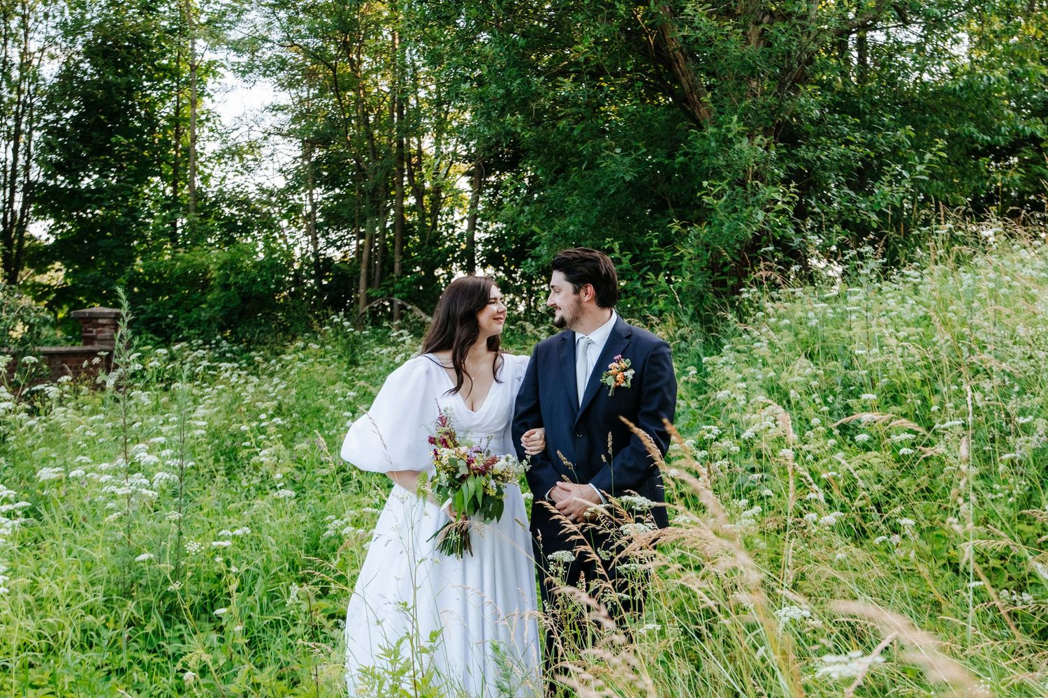 A couple walks hand in hand through a meadow of tall grass and wildflowers.