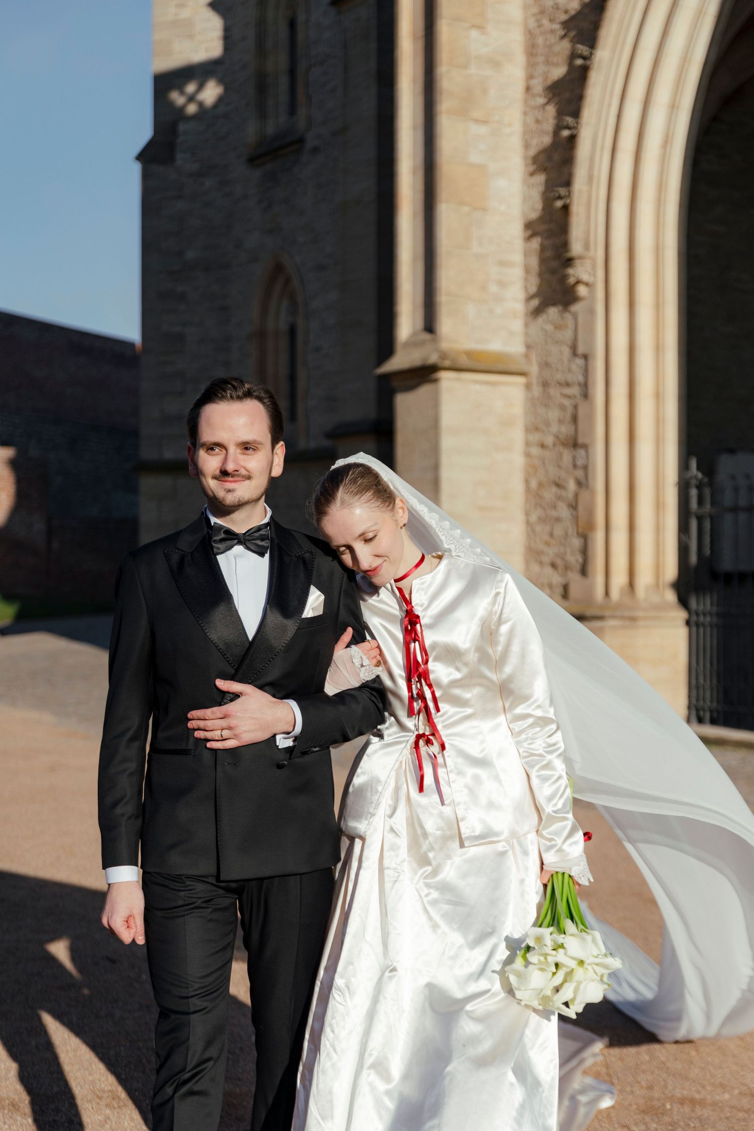 A couple in formal wedding attire share a lighthearted moment outside a stone church building in natural lighting.
