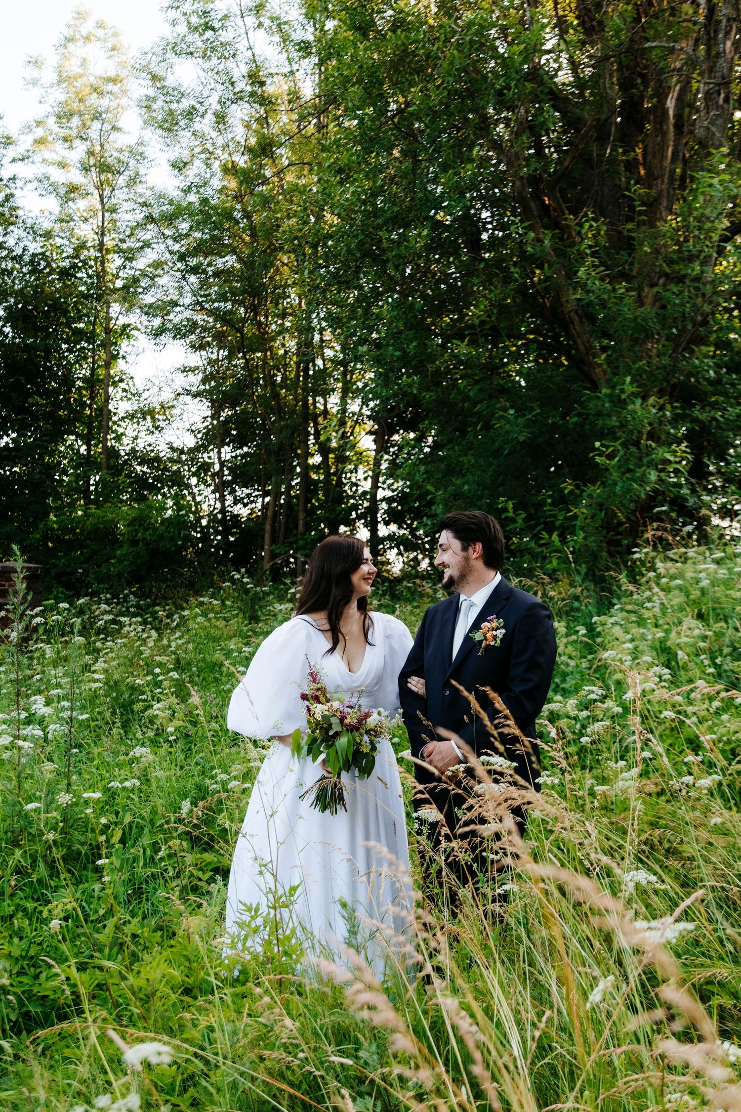 A couple in wedding attire stands together in a lush green meadow surrounded by tall trees and wildflowers.