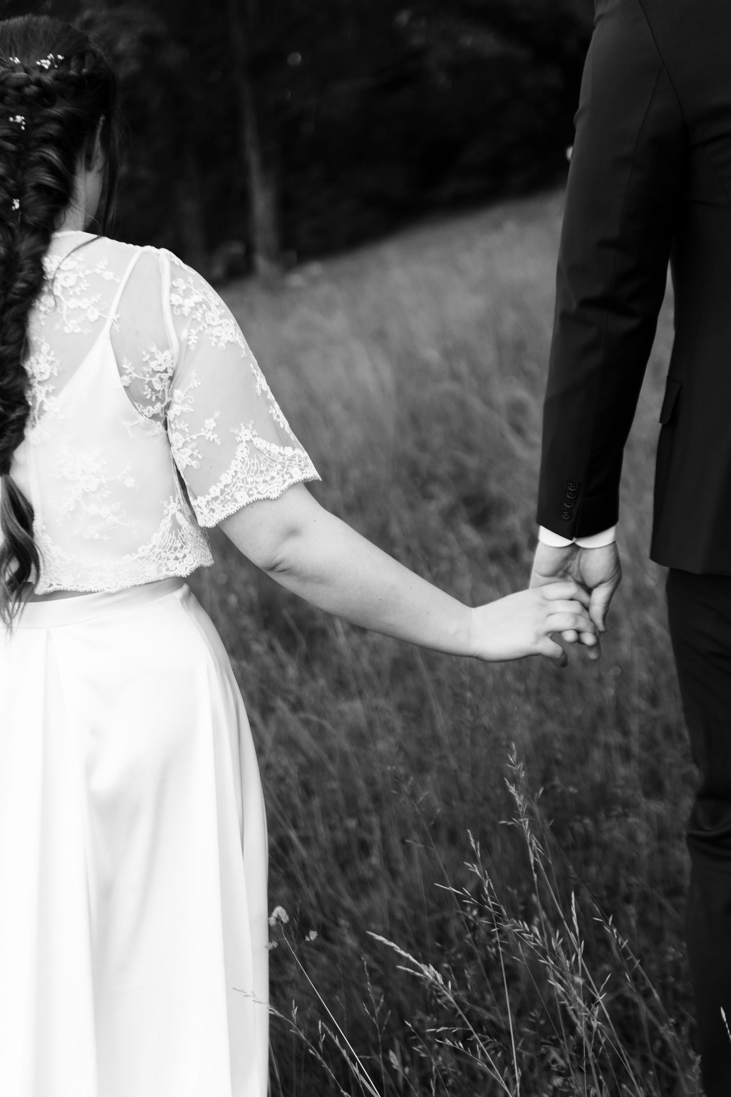 Black and white photo of bride and groom holding hands in a field, bride wearing lace wedding dress.