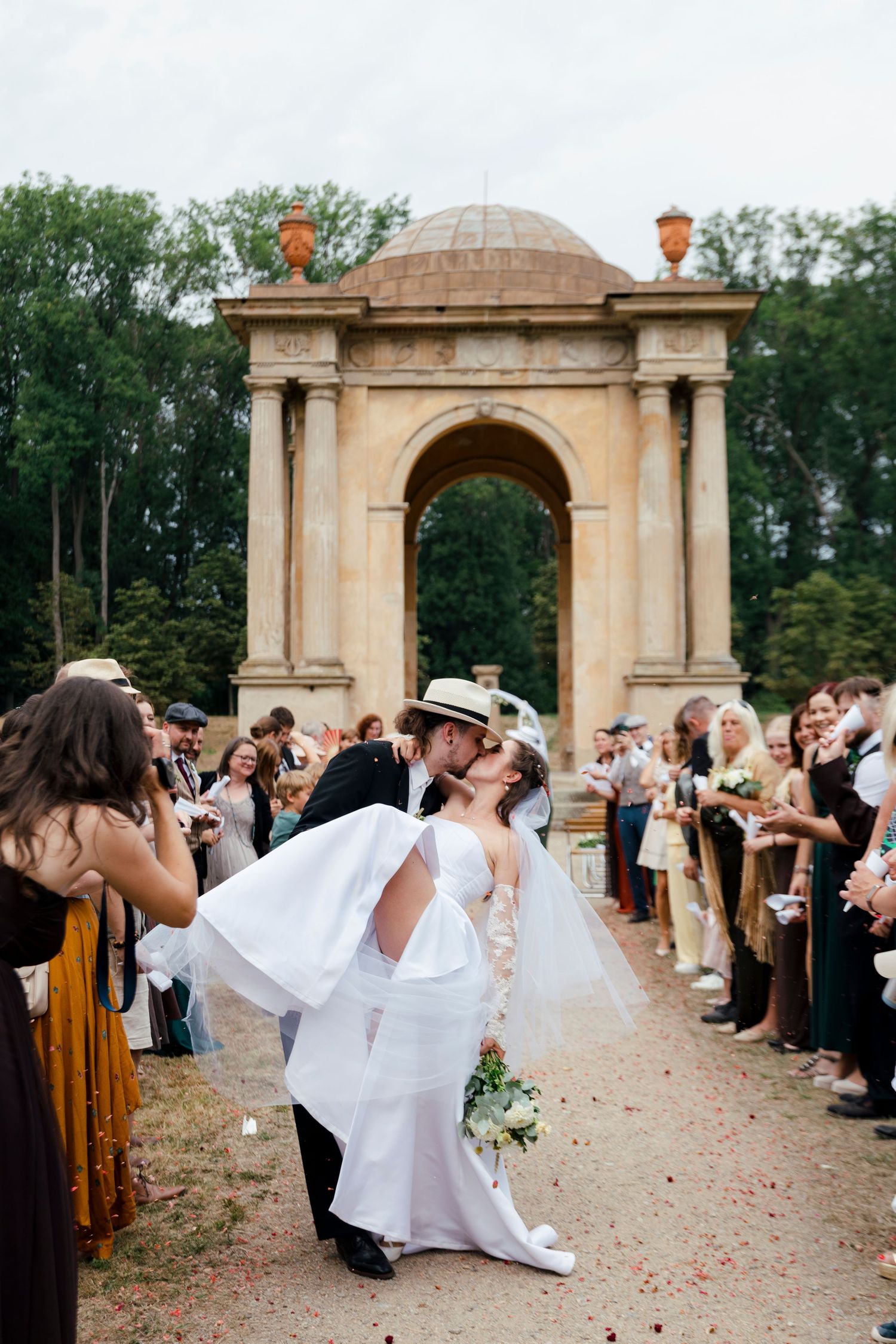 Newlyweds share a kiss on the aisle before a grand stone arch, surrounded by cheering wedding guests.