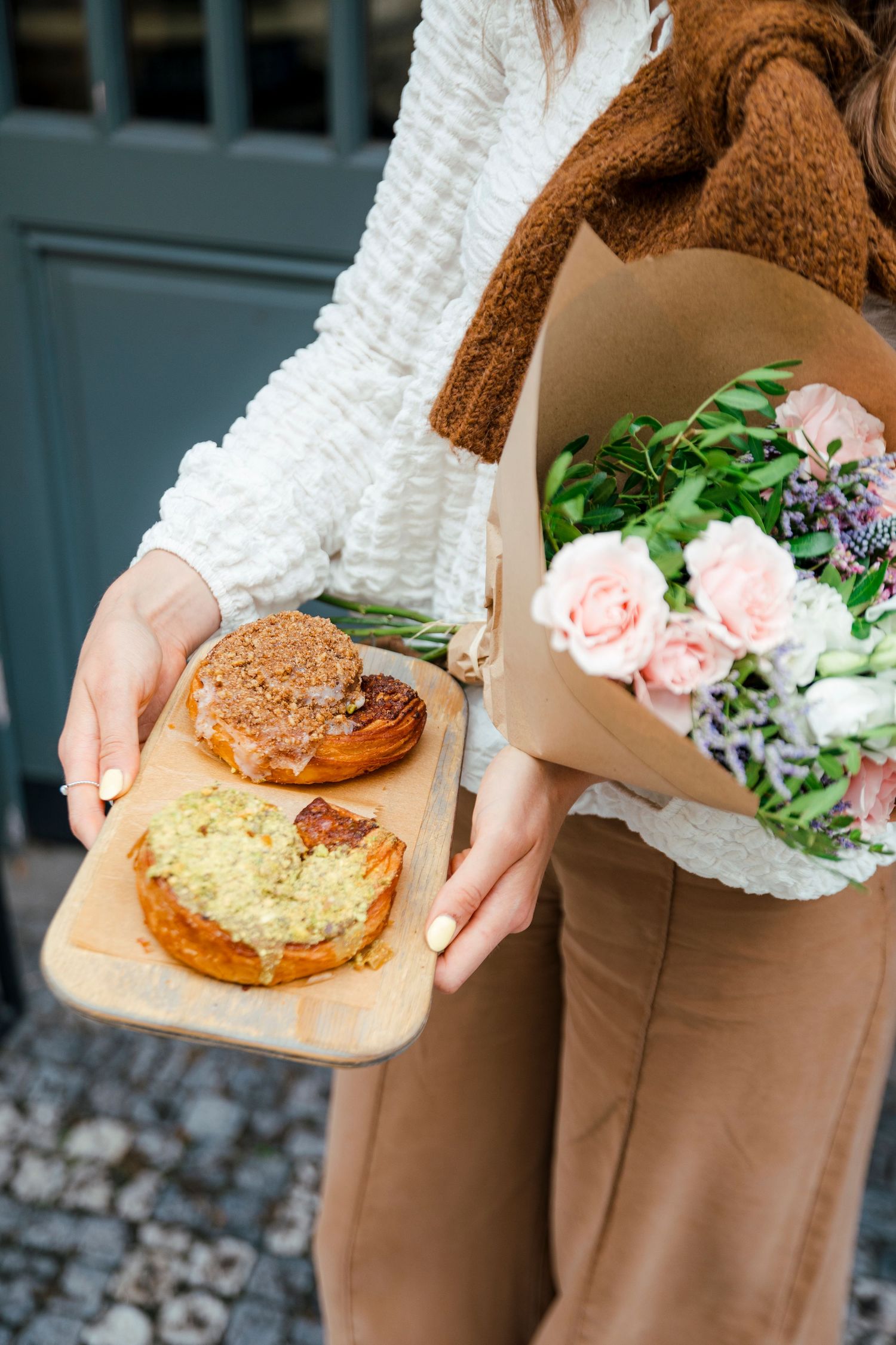 Person wearing white sweater holding pastries and flowers in brown paper wrapping.