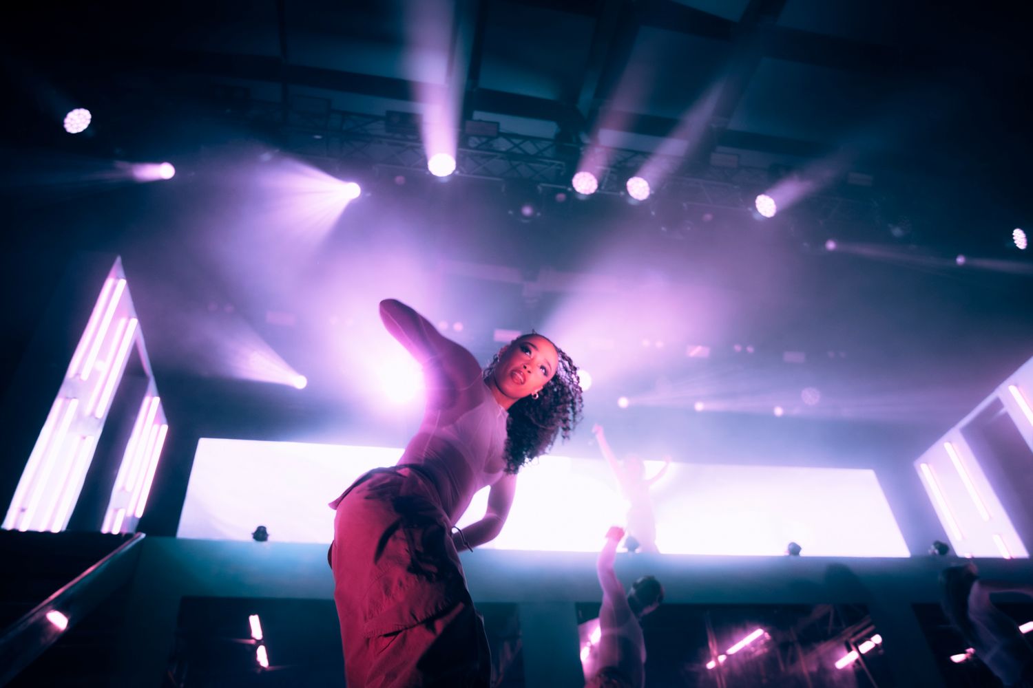 A silhouetted performer bends backwards on stage with dramatic purple lighting and beams illuminating the concert venue.