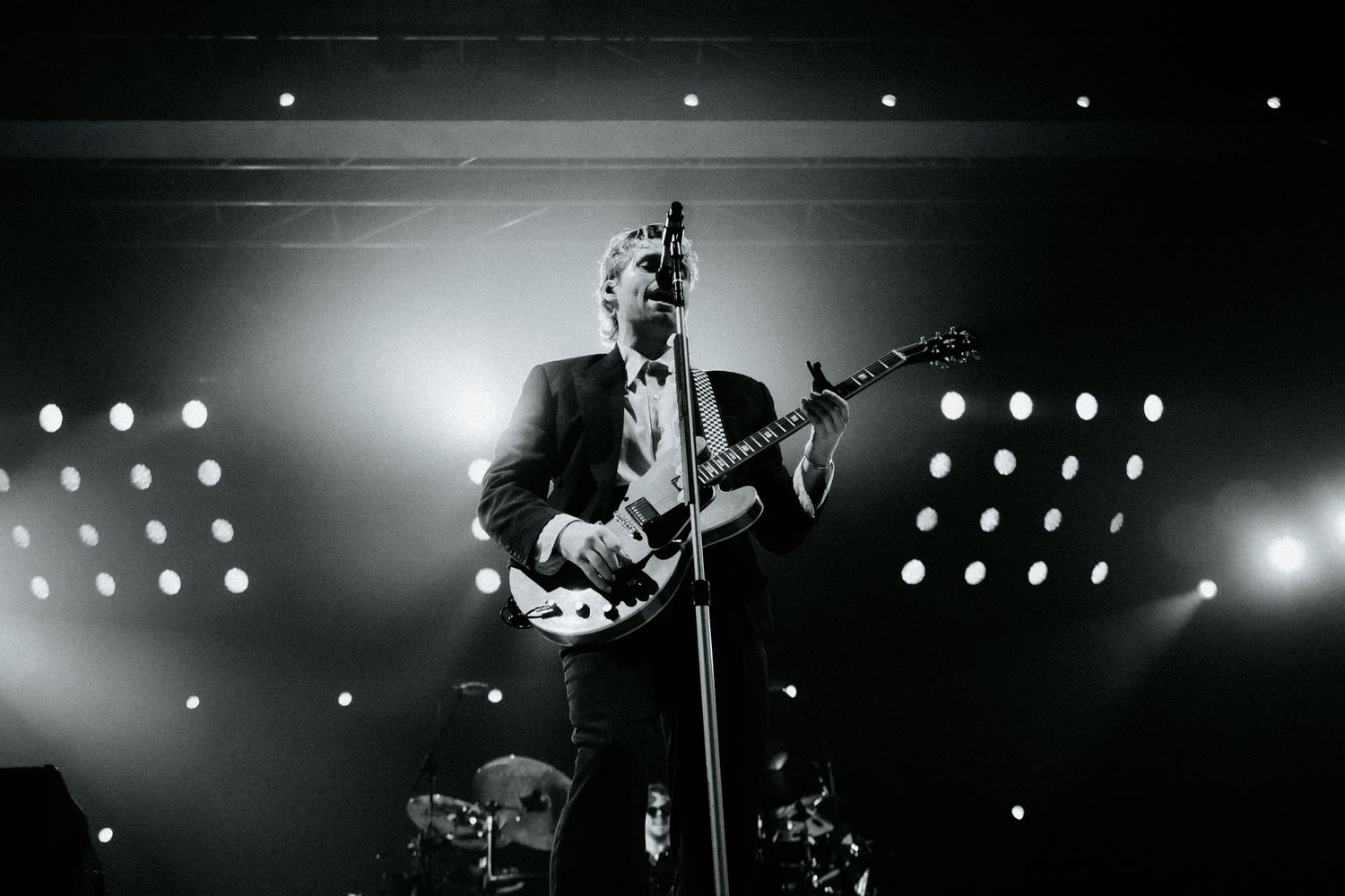 Black and white concert photo of a performer playing guitar on stage with dramatic backlighting and spotlights.