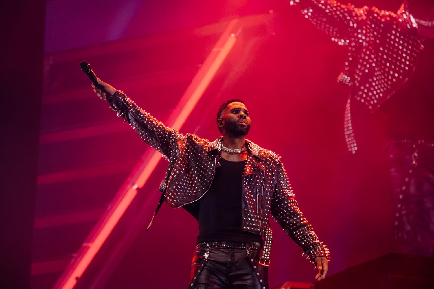 Performer in patterned jacket strikes dramatic pose on stage with red lighting and special effects in background.