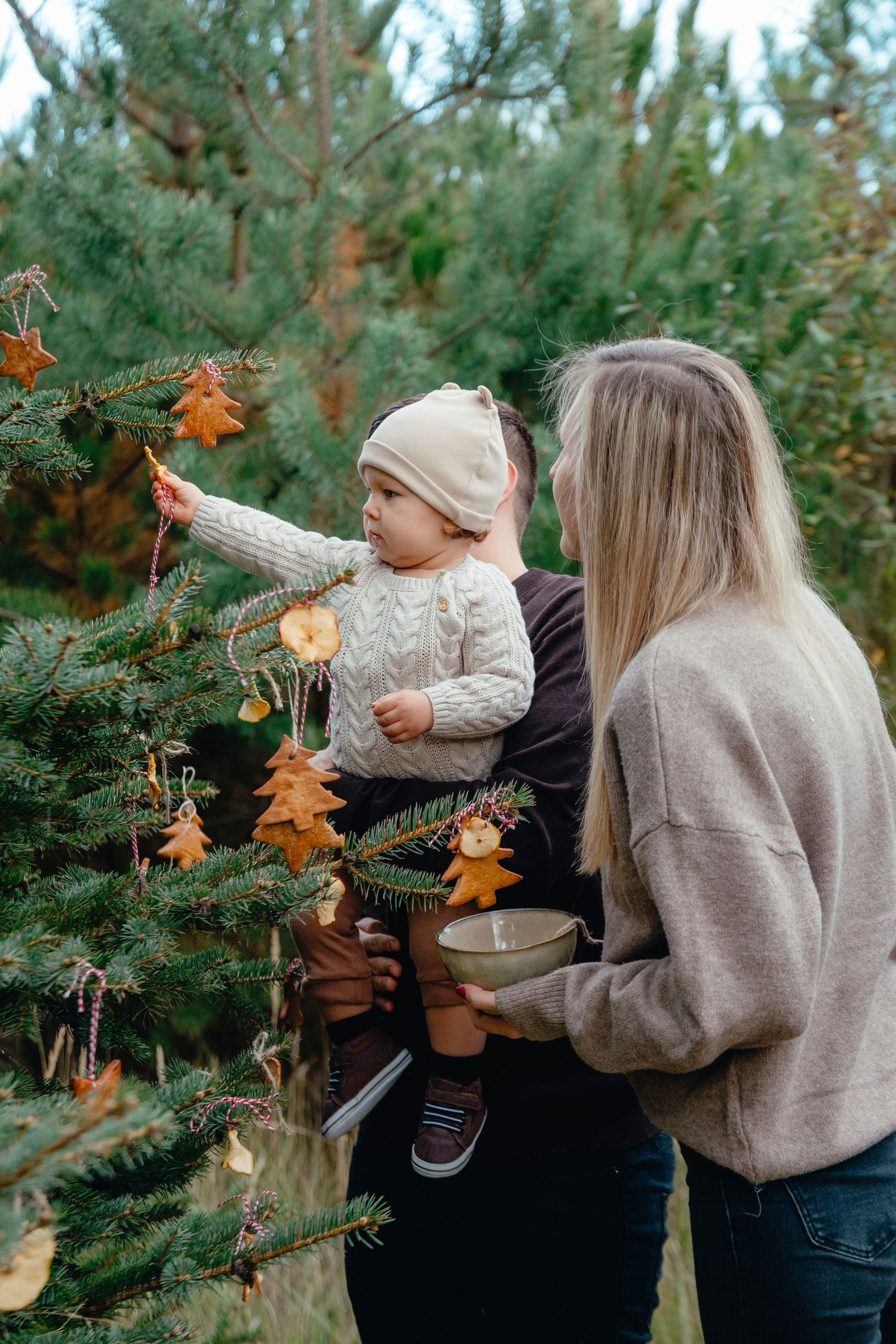 Family decorating an outdoor Christmas tree with handmade ornaments, mother holding toddler in beige hat and knit sweater.