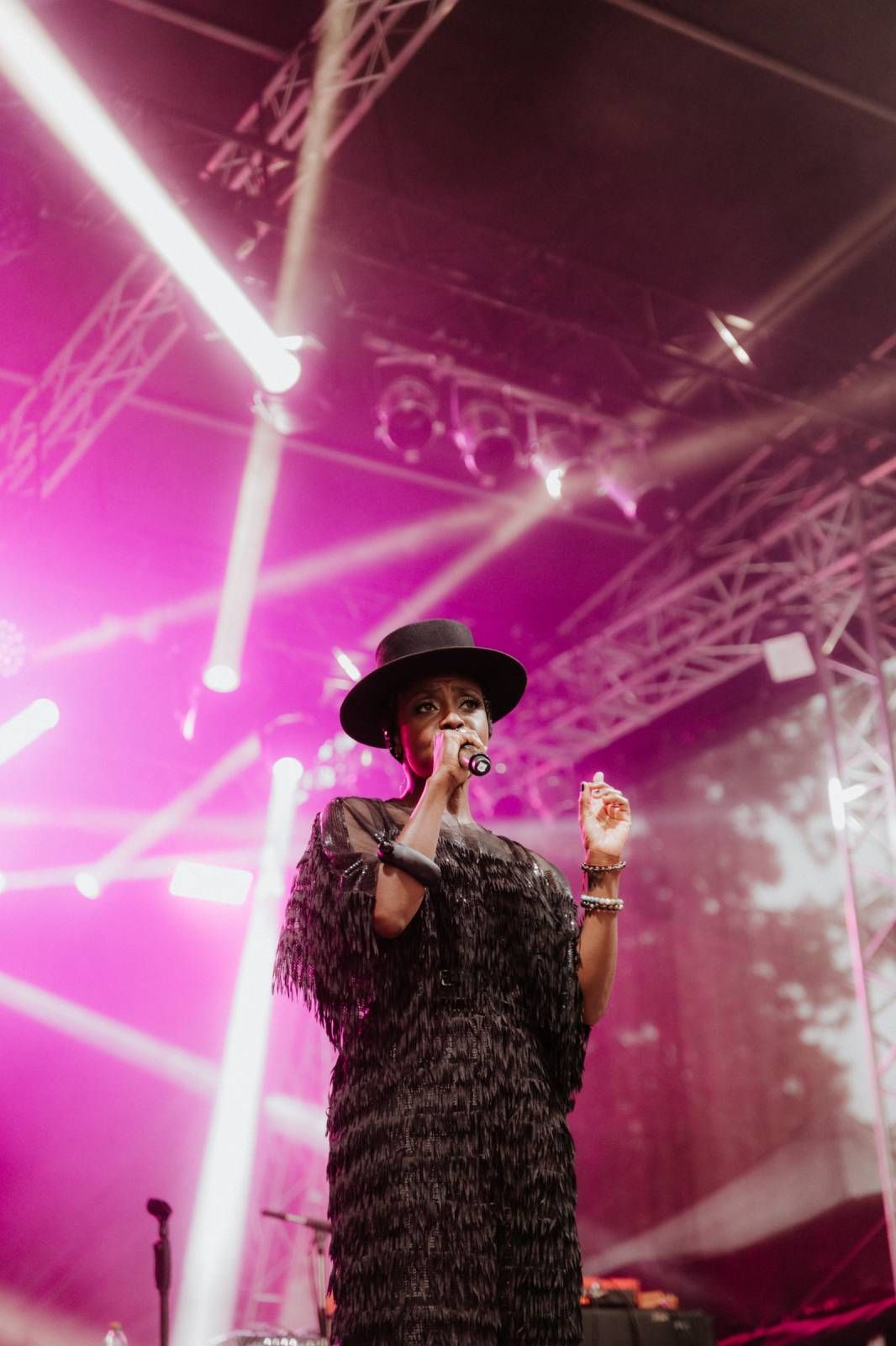 A performer in a black feathered outfit and hat sings on stage with dramatic pink lighting and fog effects.