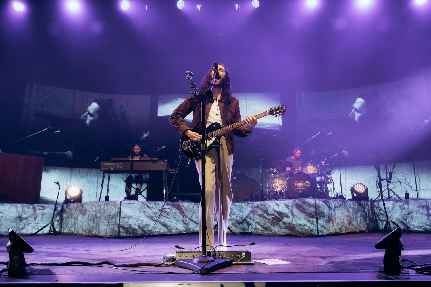 A performer stands on stage bathed in purple lighting while large video screens display nature scenes in the background.