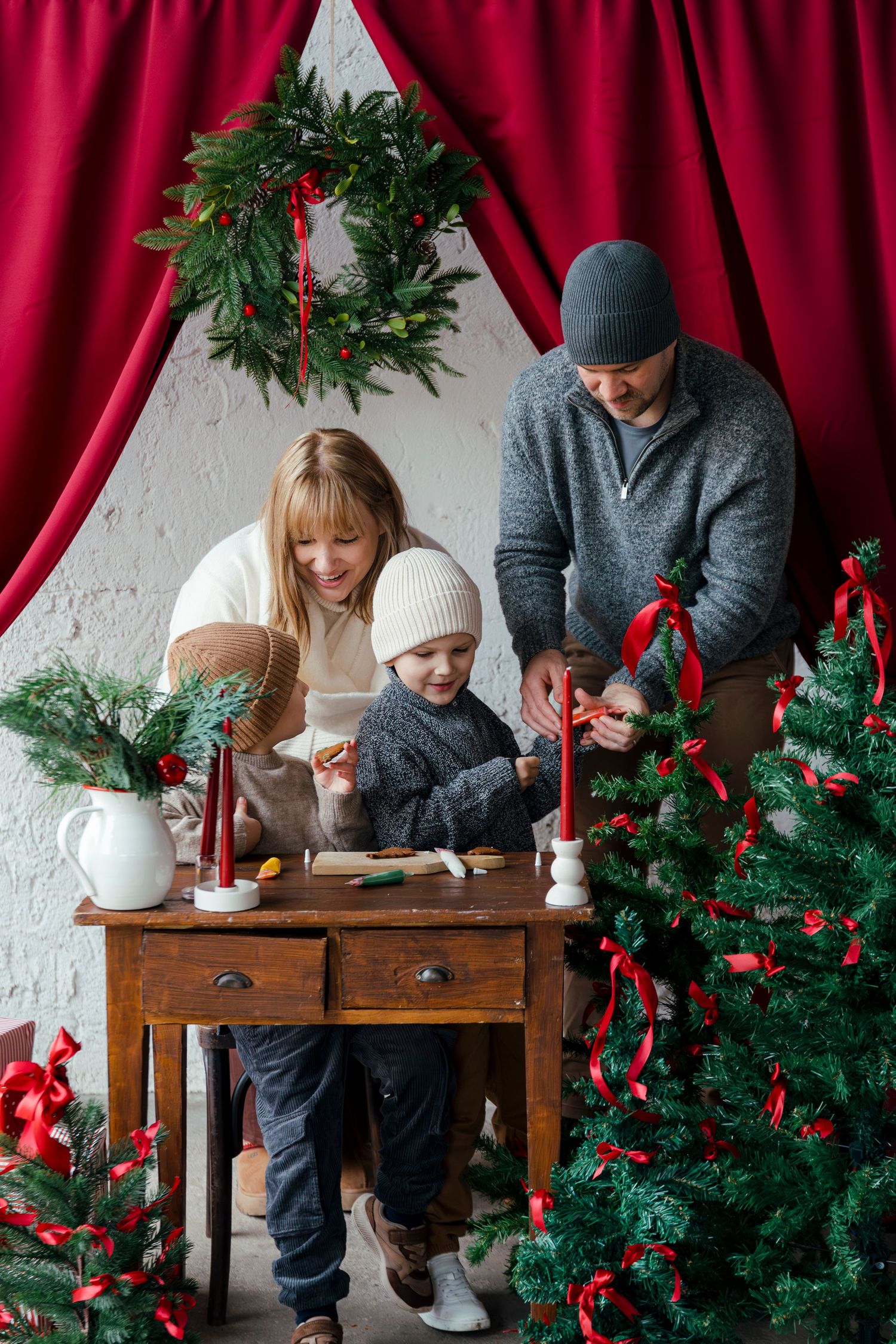 Family decorating for Christmas near a tree, wreath, and red curtains, with a child at a wooden table.