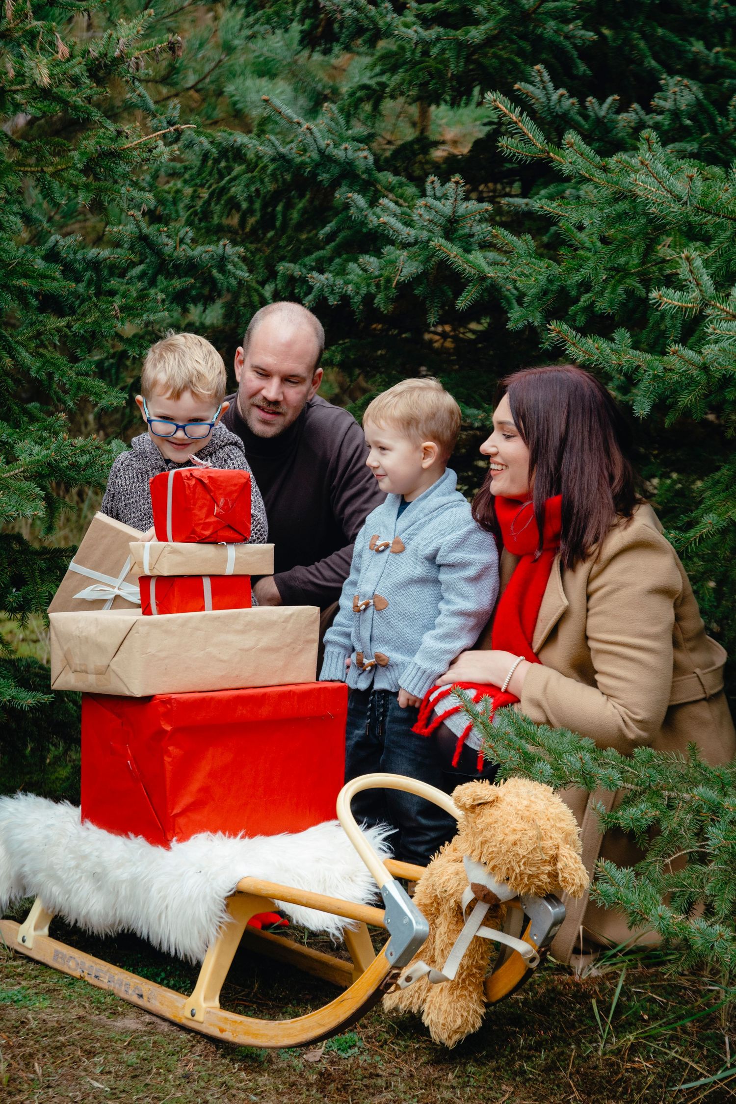 Family with two young boys opening Christmas gifts on a sled outdoors near evergreen trees.