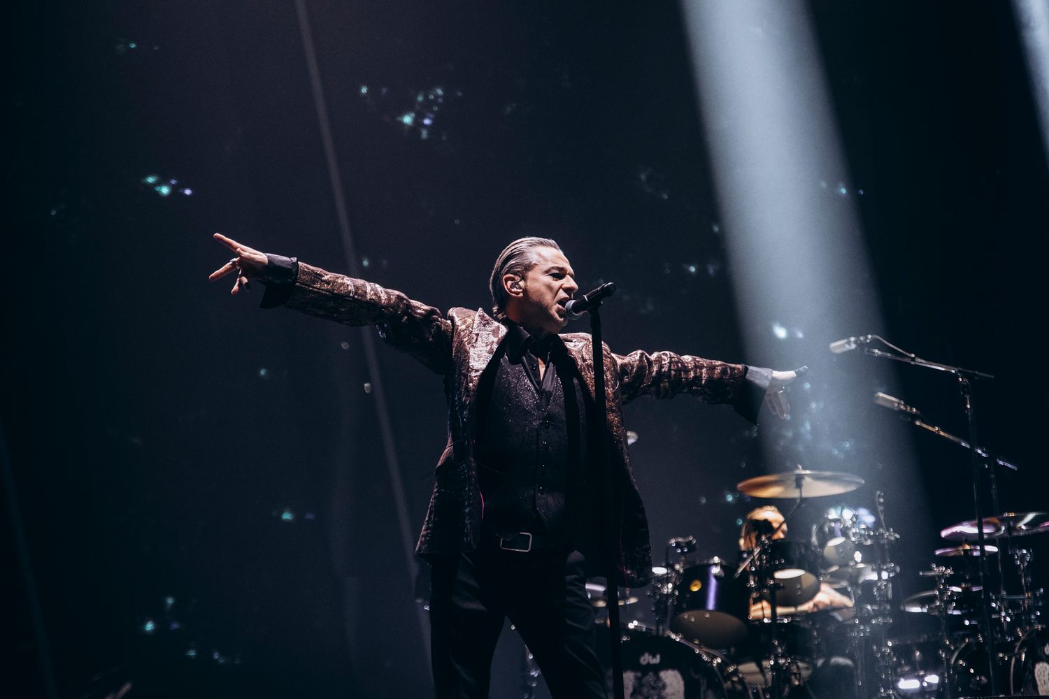 Concert performer on stage with arms outstretched in dramatic pose against dark background with spotlights.
