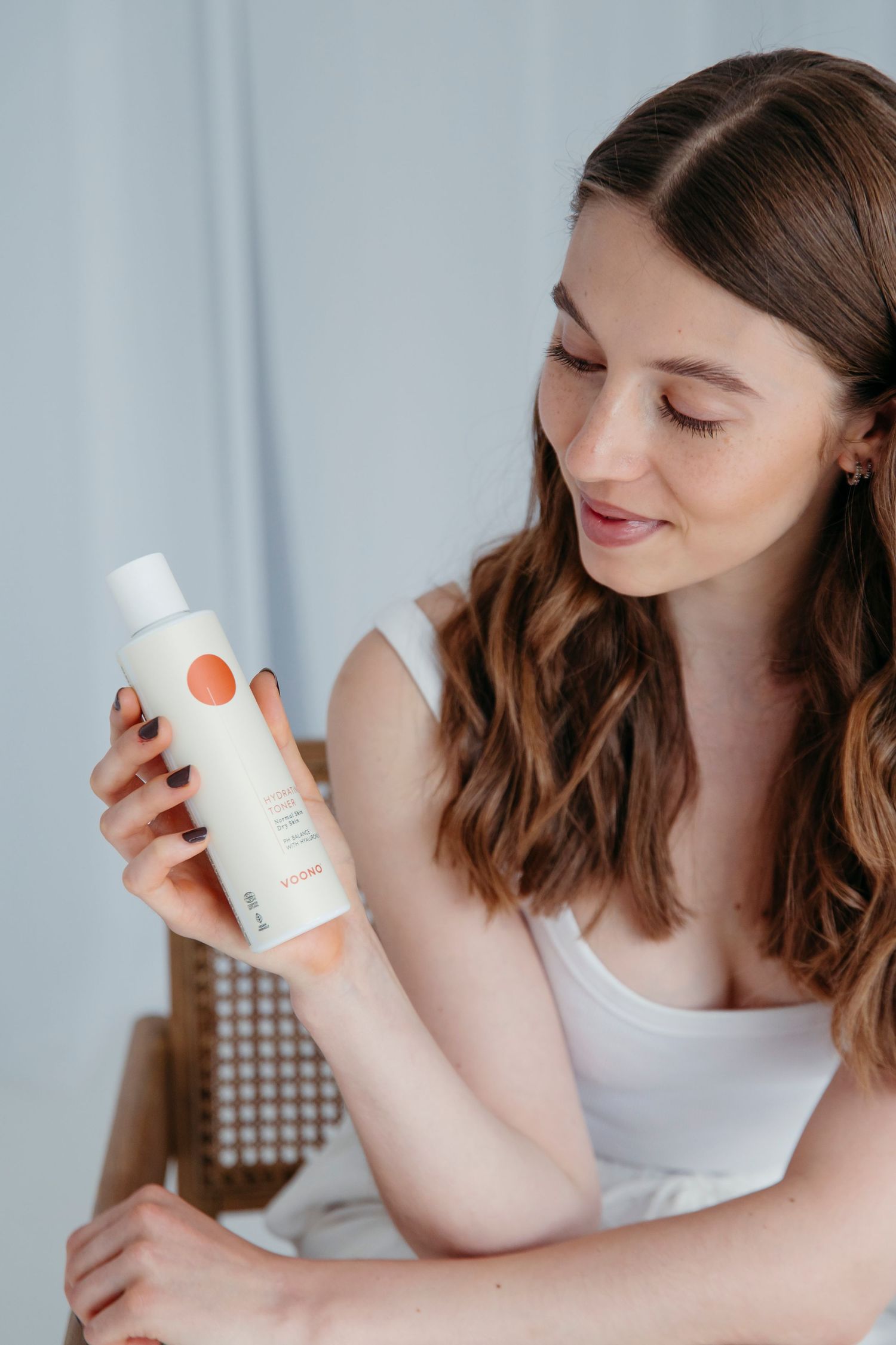 Woman examining skincare product bottle against light background.