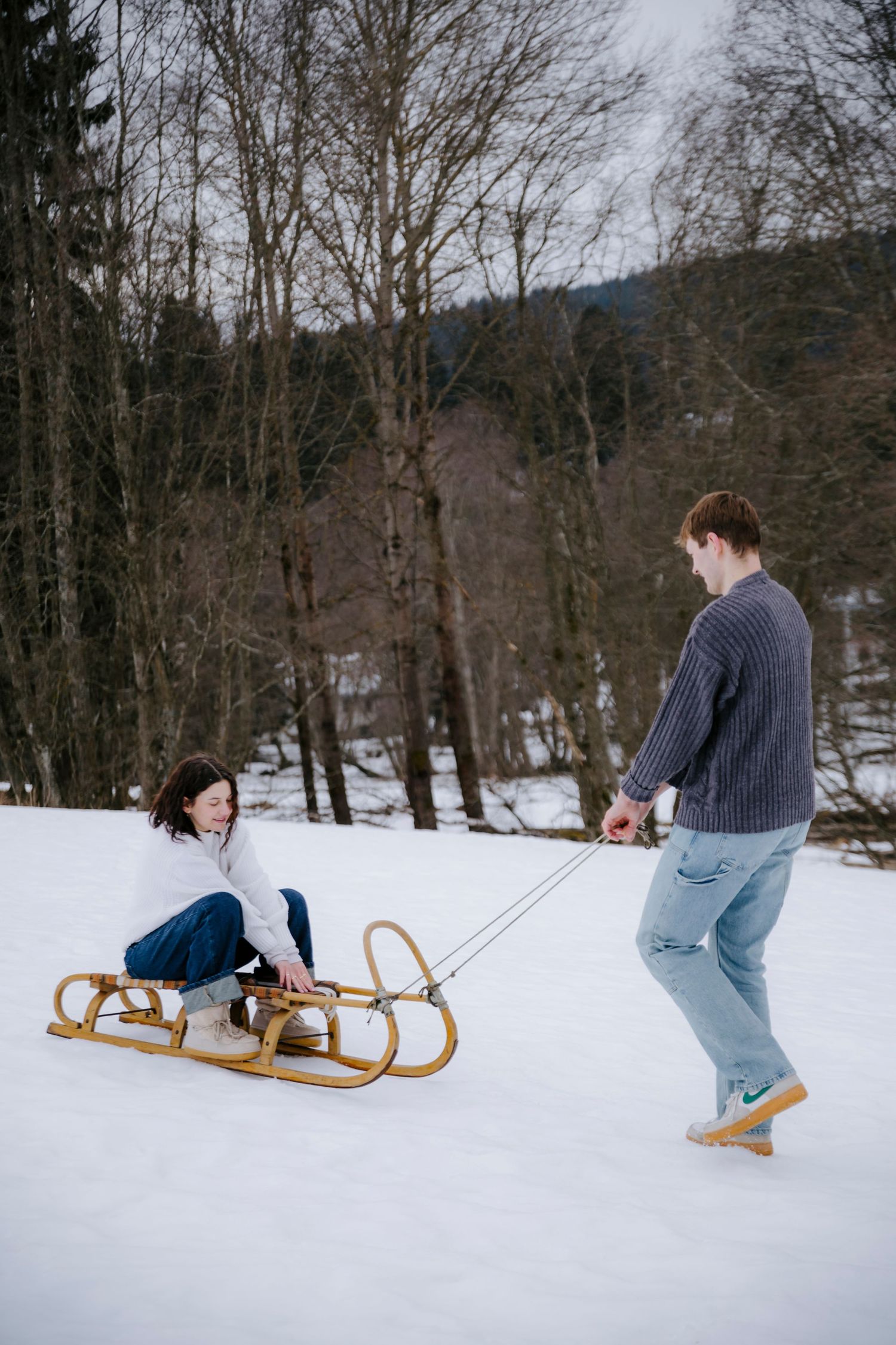 Person pulling another on a wooden sled through snowy winter landscape.
