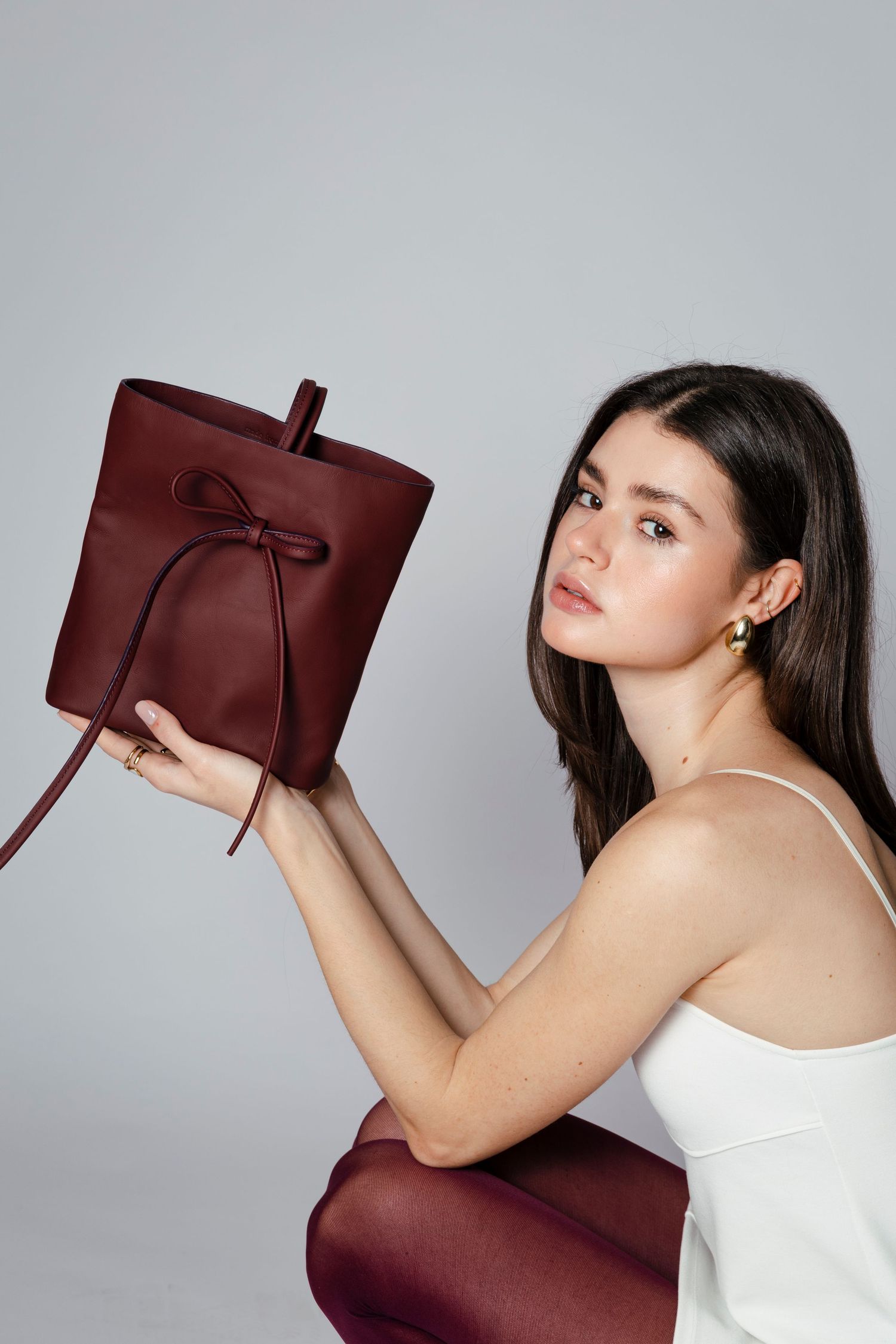 Fashion model in white tank top holding burgundy leather bucket bag against gray background.