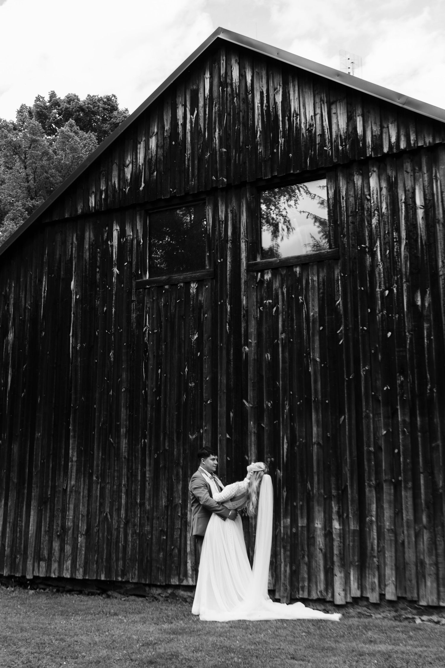 Black and white photo of bride and groom embracing beside a rustic wooden barn on their wedding day.