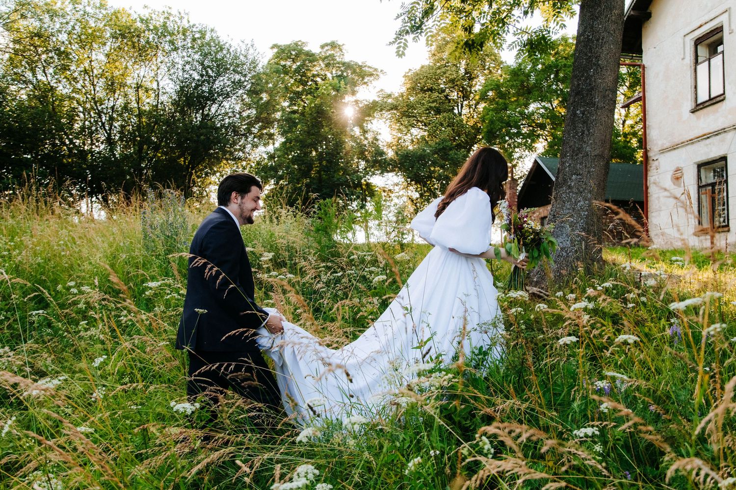 A romantic couple poses together in a lush green field with wildflowers and trees.