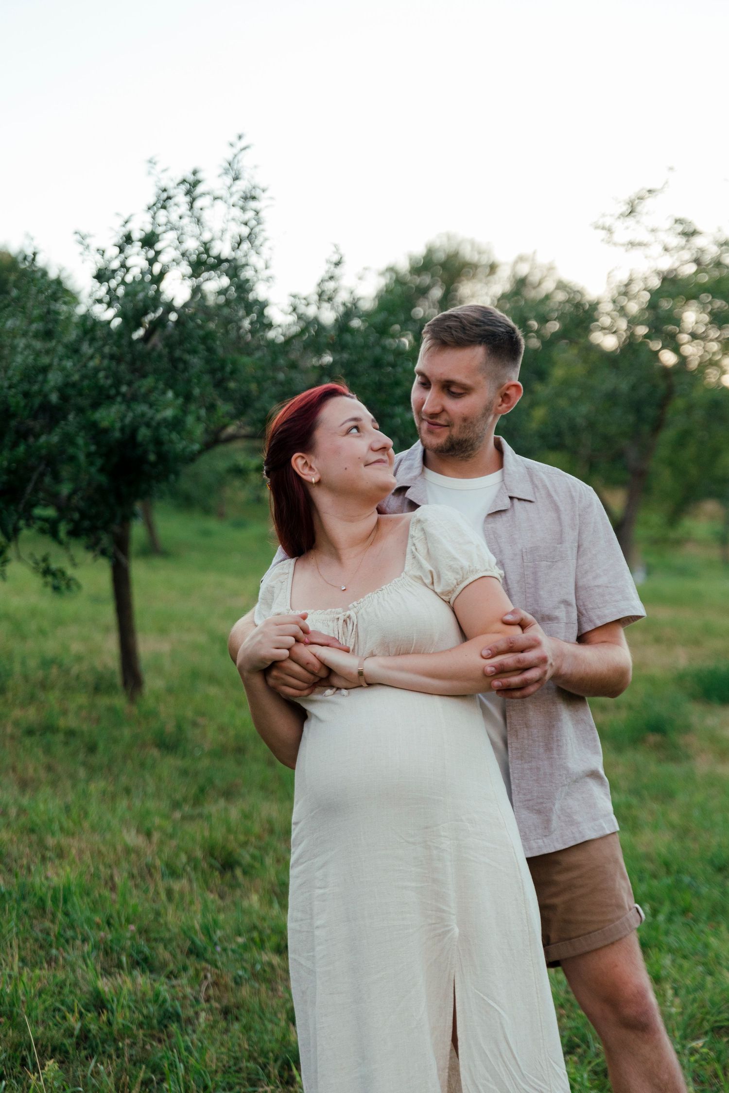 Pregnant woman in white dress embraced by man in garden setting, couple smiling outdoors among green trees.