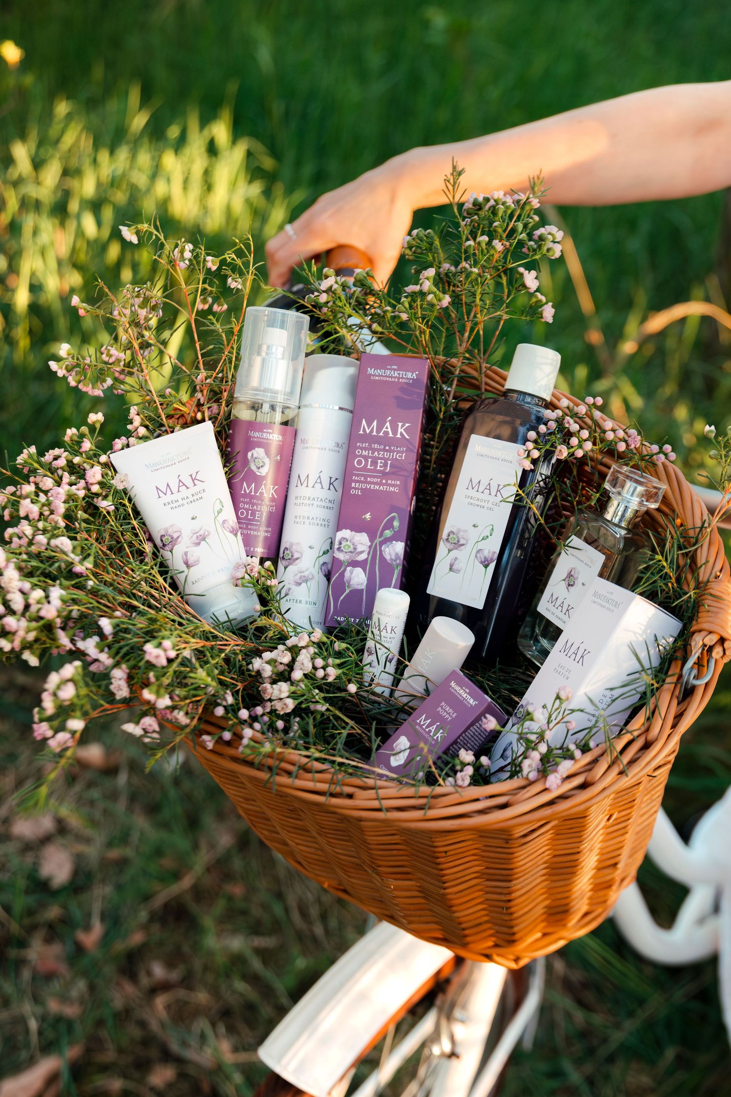 A wicker basket filled with MAK brand skincare products and pink wildflowers, held by a hand outdoors.