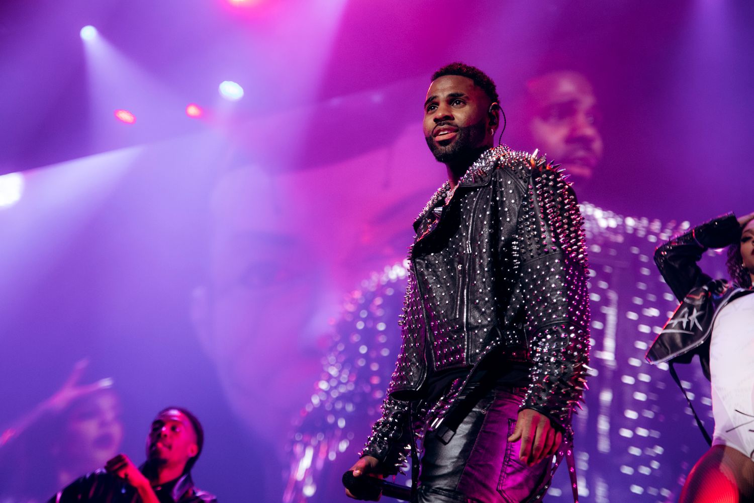 A performer in a studded black jacket dances on stage with vibrant purple and pink lighting effects in the background.