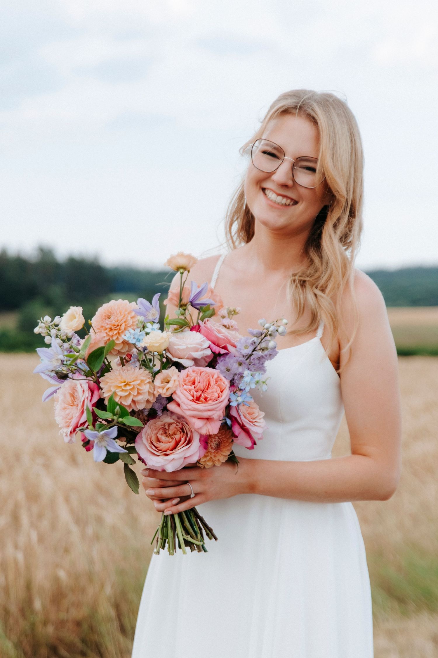 Bride holding vibrant bouquet of pink, peach and purple flowers in wheat field.