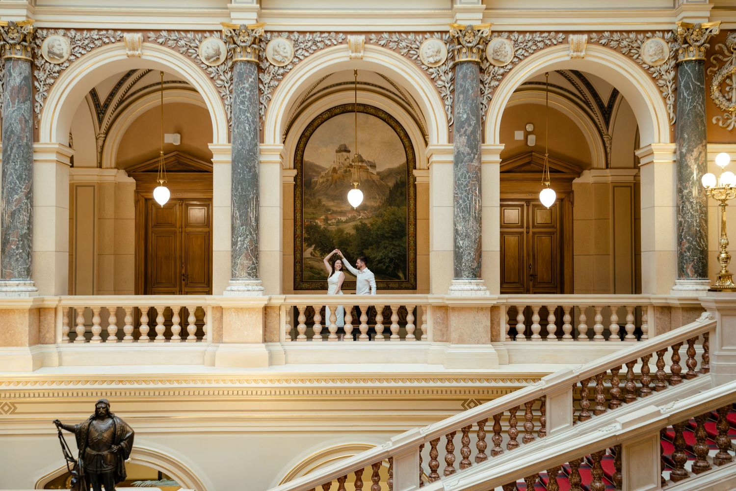 Elegant couple poses in arched balcony of historic building with marble columns and ornate architecture.