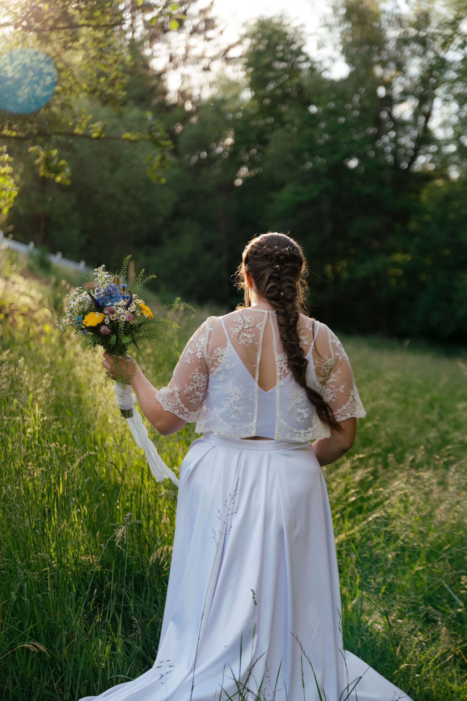Bride in white lace top and skirt holds wildflower bouquet in sunlit green meadow, viewed from behind.