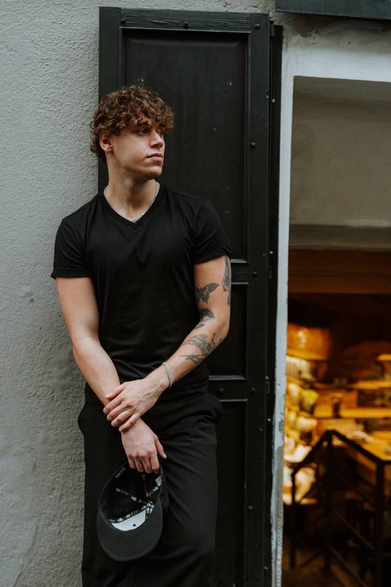 Person in black t-shirt leans against white wall near dark doorway with warm lighting visible inside.